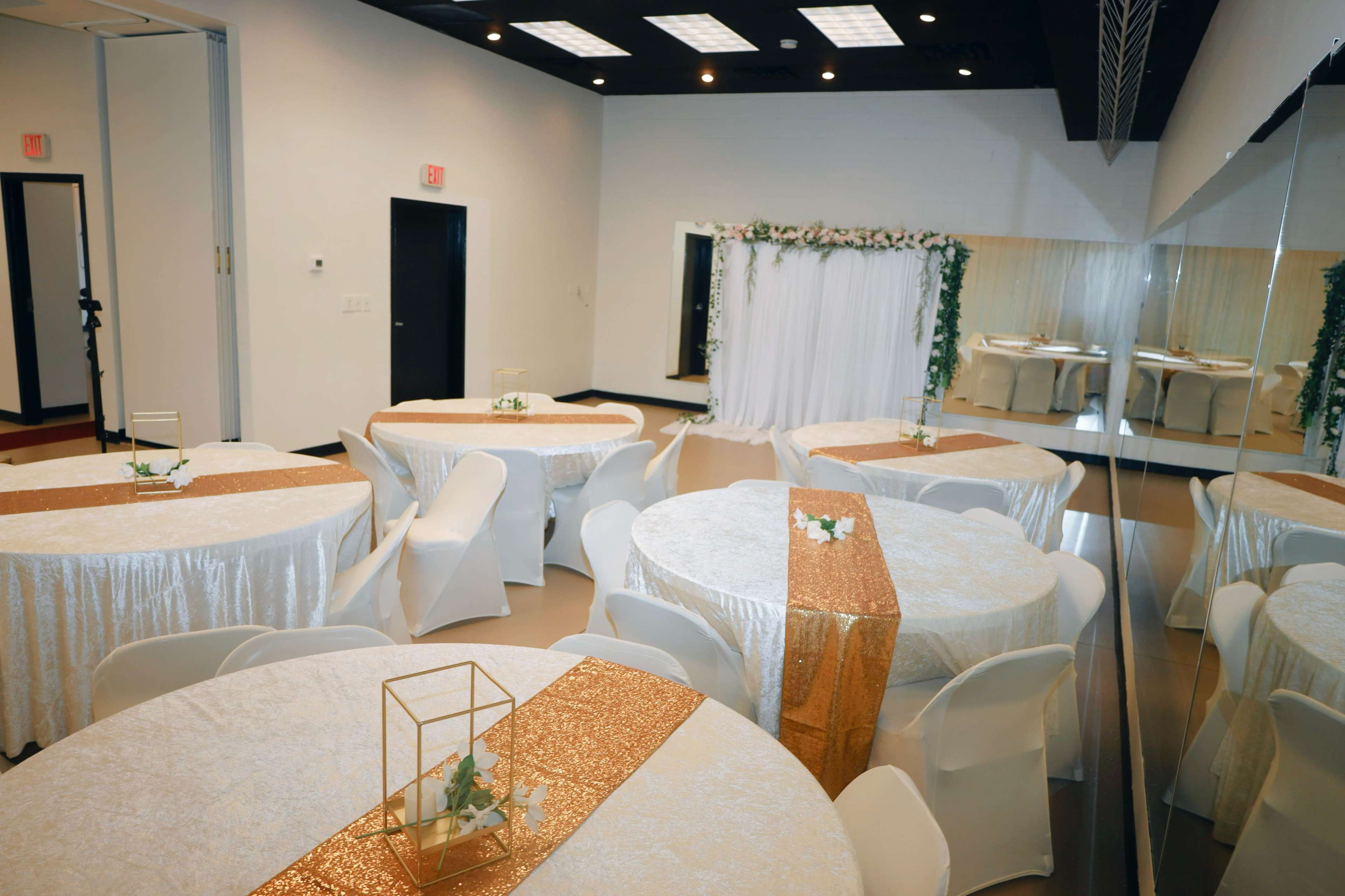 An elegantly arranged event space with round tables covered in white tablecloths and gold runners, surrounded by white chairs and a decorative backdrop.