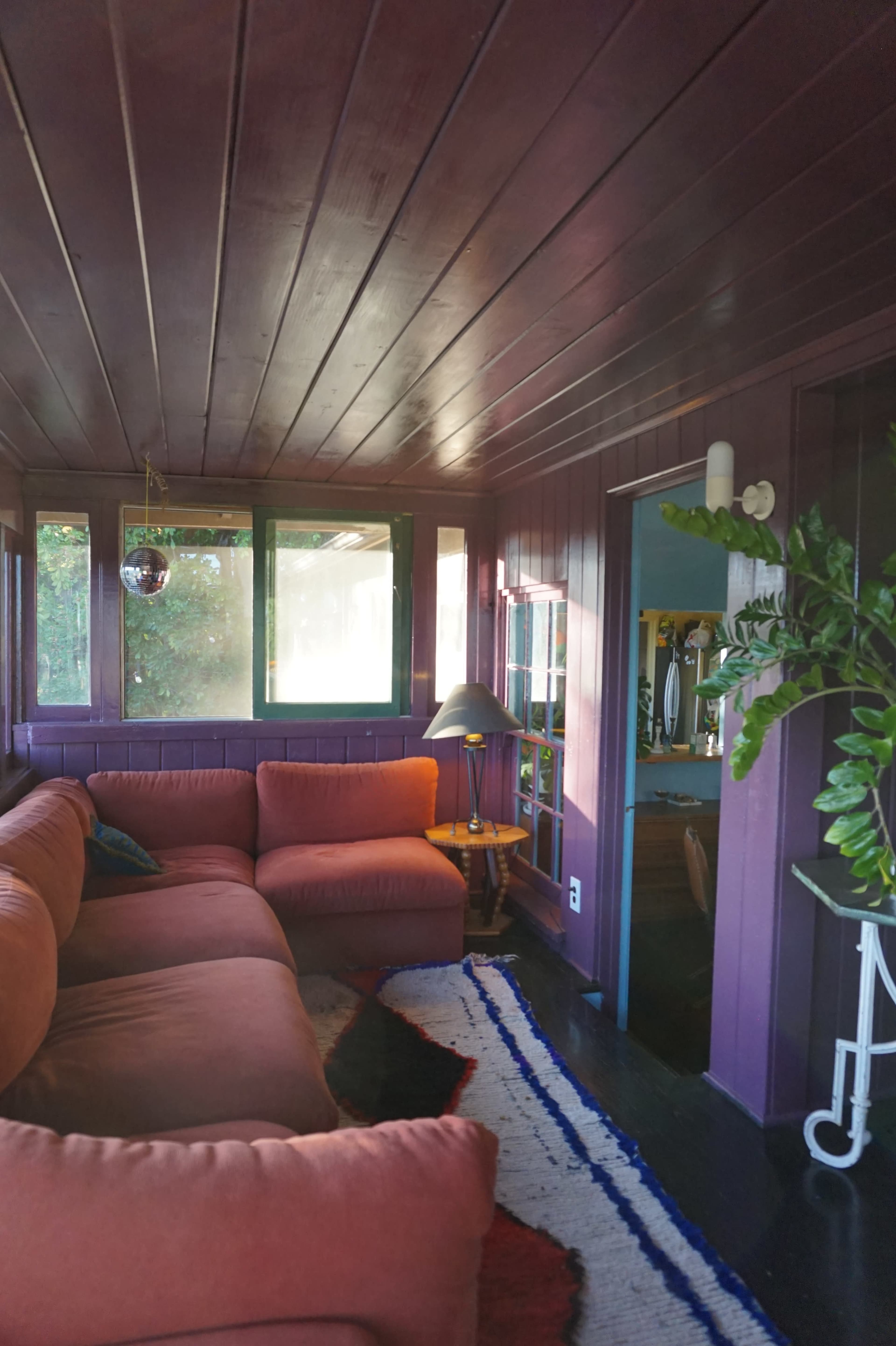 The image shows a living room with a large pink couch, a lamp, and green plants, all set against a purple wooden wall and large windows.
