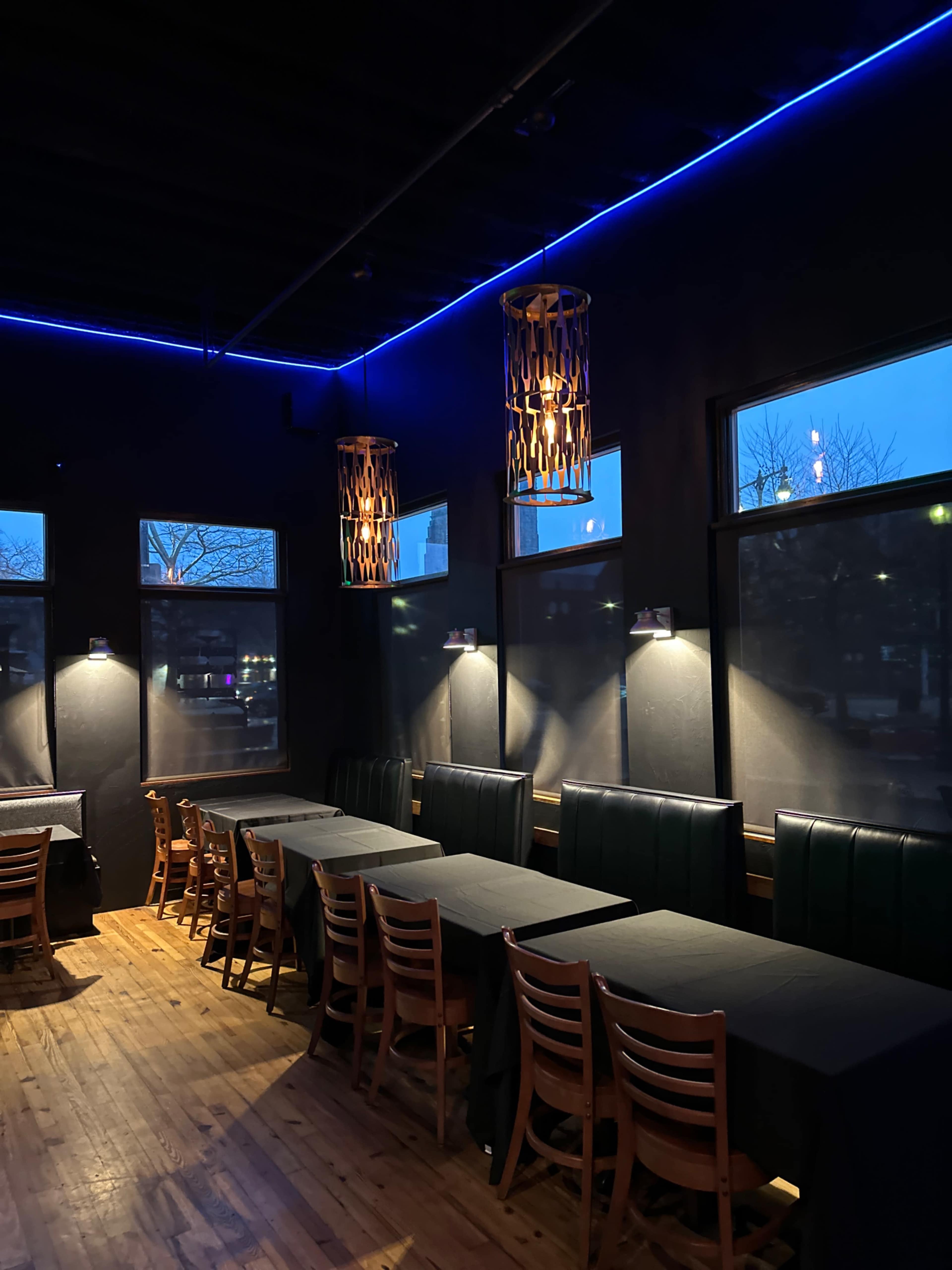 The interior of a restaurant features wooden tables with black tablecloths, illuminated by pendant lights and blue LED accents along the ceiling.