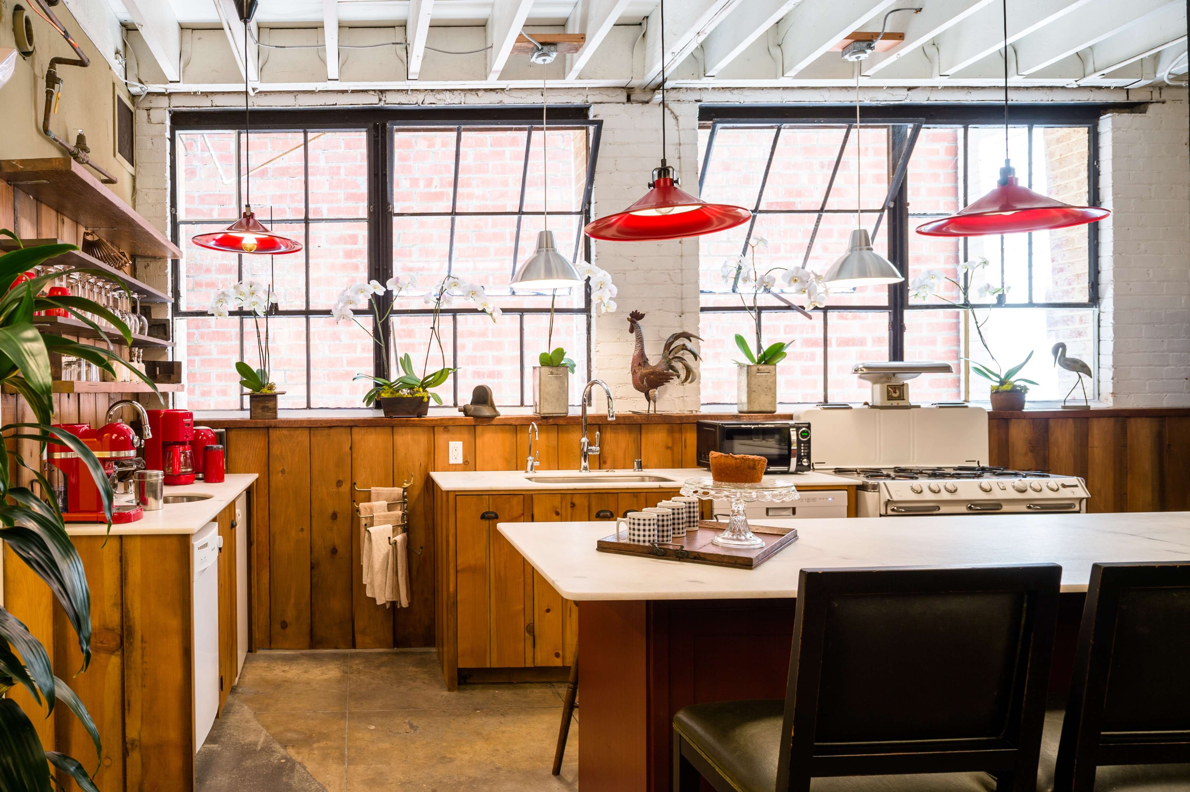 The kitchen features wooden cabinetry, a large island, red pendant lights, and large windows overlooking a brick wall.