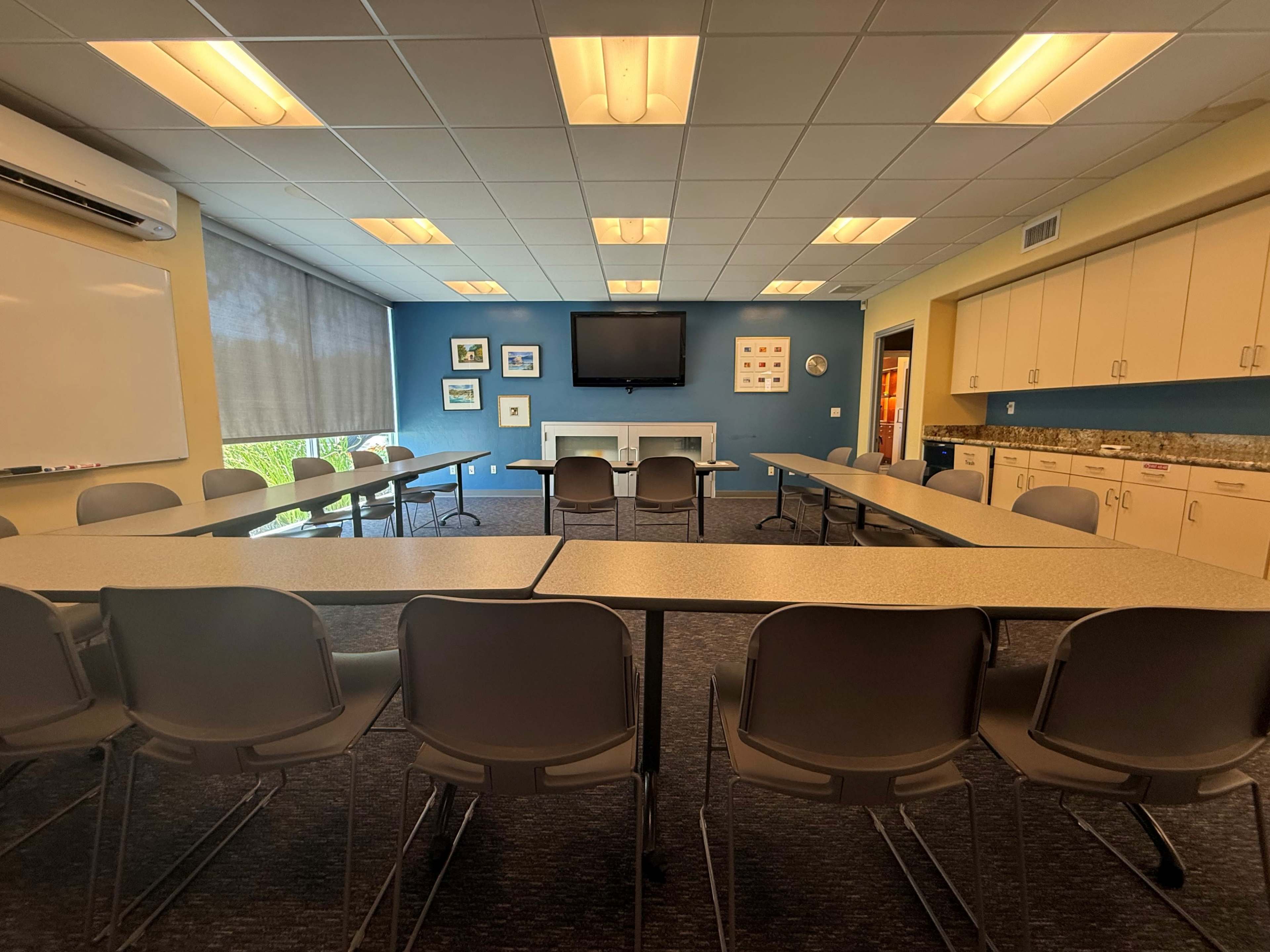 The image shows a meeting room with multiple tables arranged in a U-shape, surrounded by gray chairs, and featuring a TV screen on the wall.