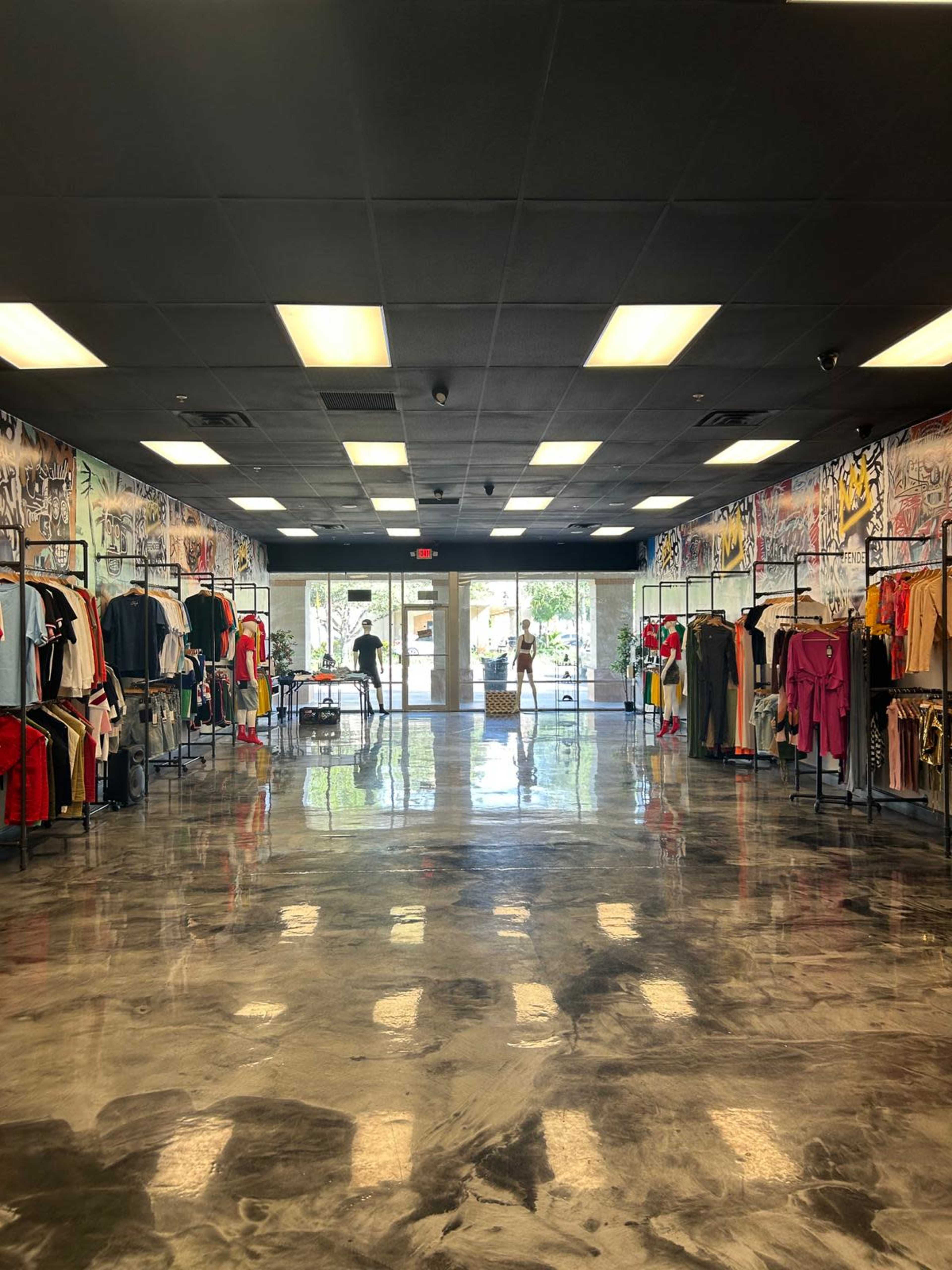 A clothing store features various apparel hanging on the walls, with polished concrete floors reflecting the light from large windows at the front.