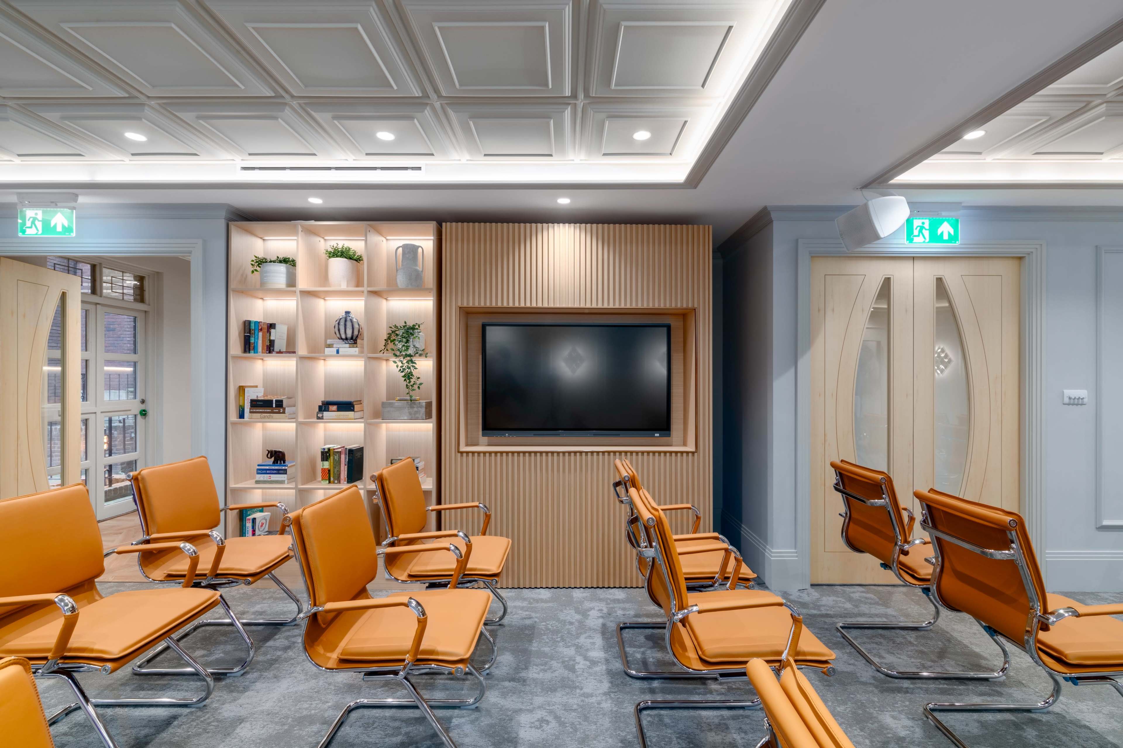 A waiting room features several orange chairs arranged in rows, with a wooden accent wall that includes a television and decorative shelves in the background.