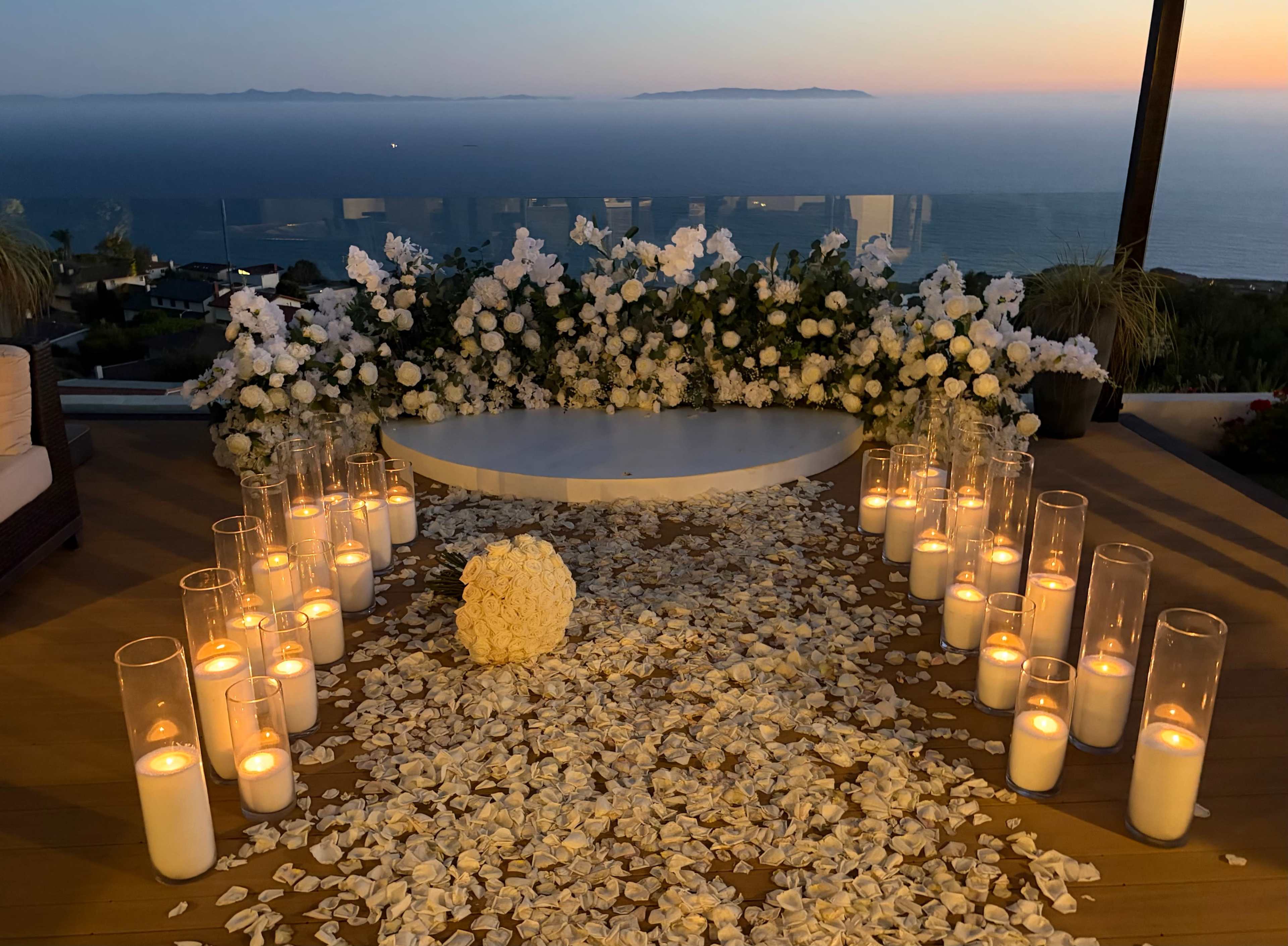 The image shows a wedding ceremony setup with a row of candles and a floral arrangement overlooking the ocean at sunset.