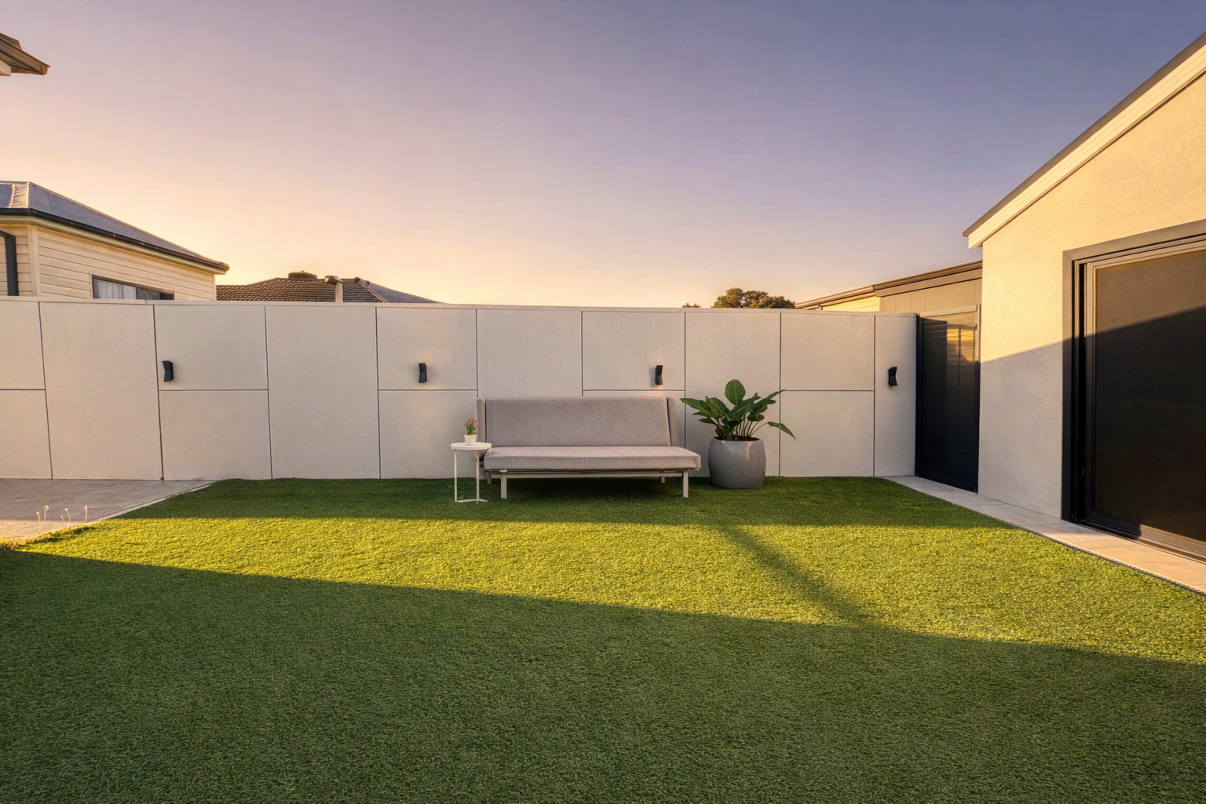The image shows a modern backyard with a gray couch on artificial grass, surrounded by a concrete wall and a small potted plant.