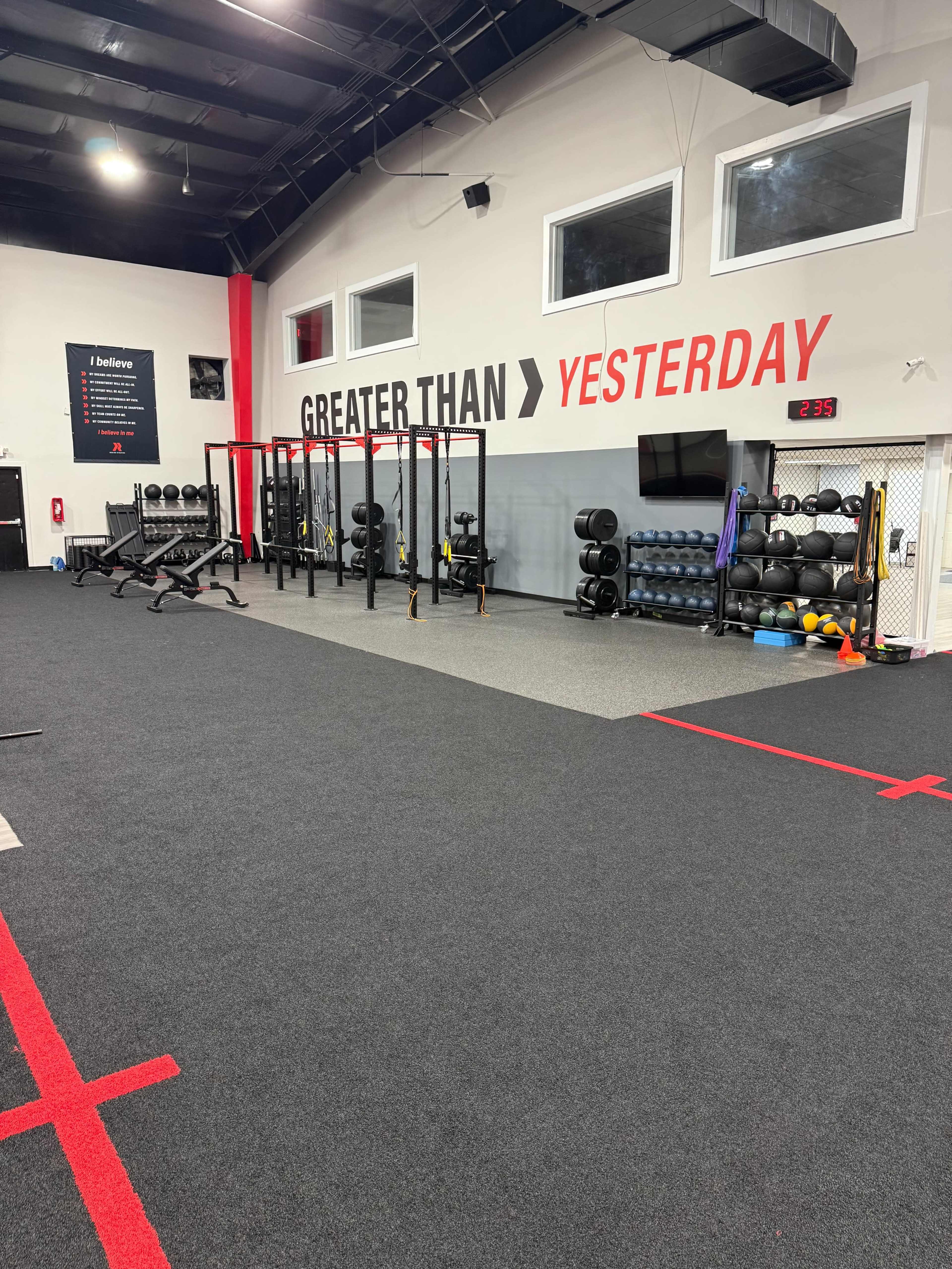 The image shows a spacious gym interior with various fitness equipment organized along the walls, including weights and exercise mats, with a motivational phrase displayed prominently on the back wall.