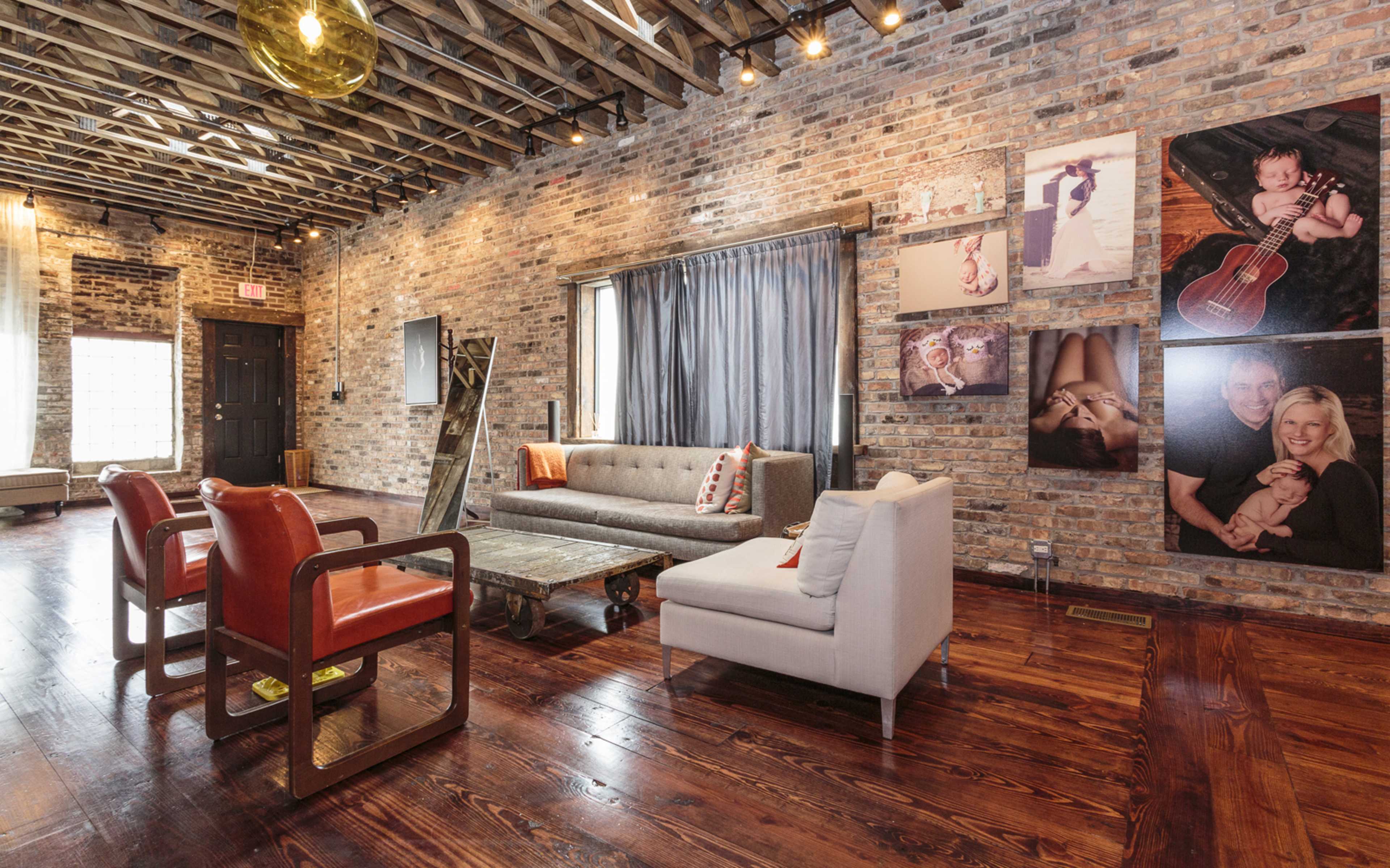 A modern living room with exposed brick walls, hardwood floors, and various seating arrangements, including a gray sofa and orange-accented chairs.