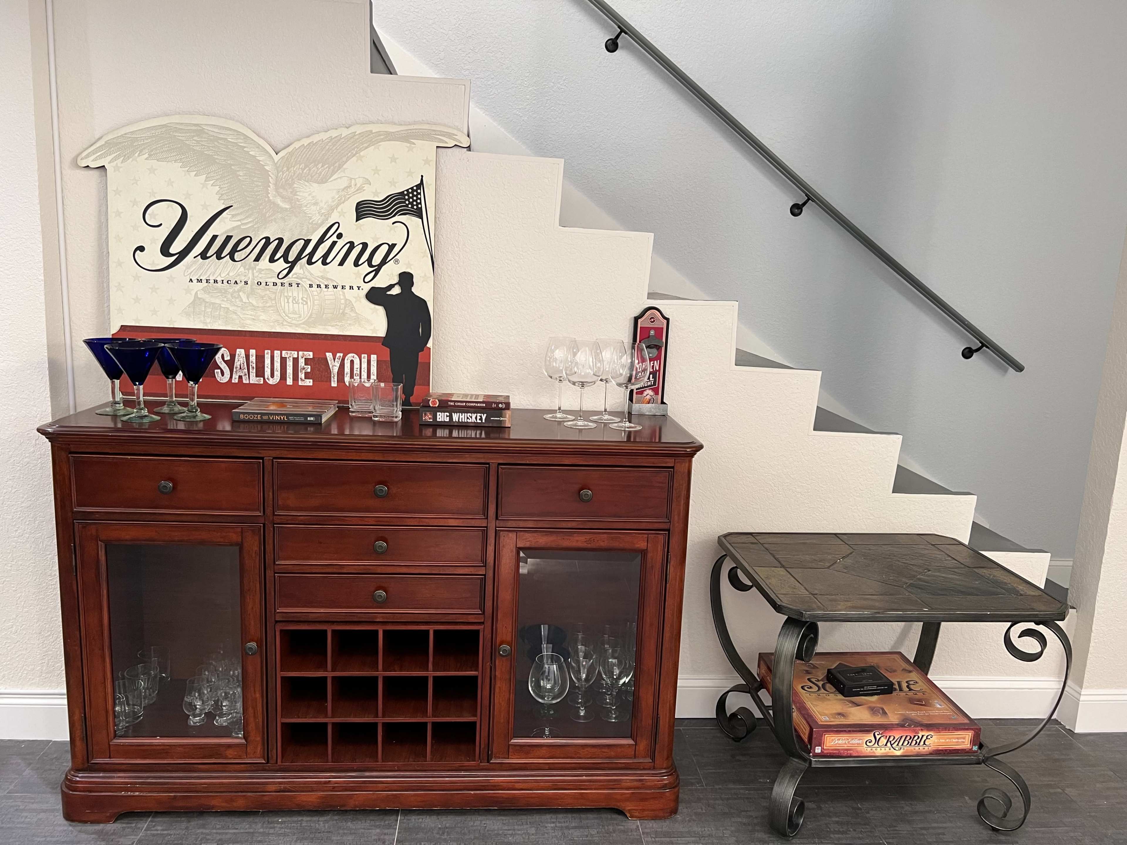 A wooden sideboard with glassware and a decorative sign is positioned against a wall near a staircase.