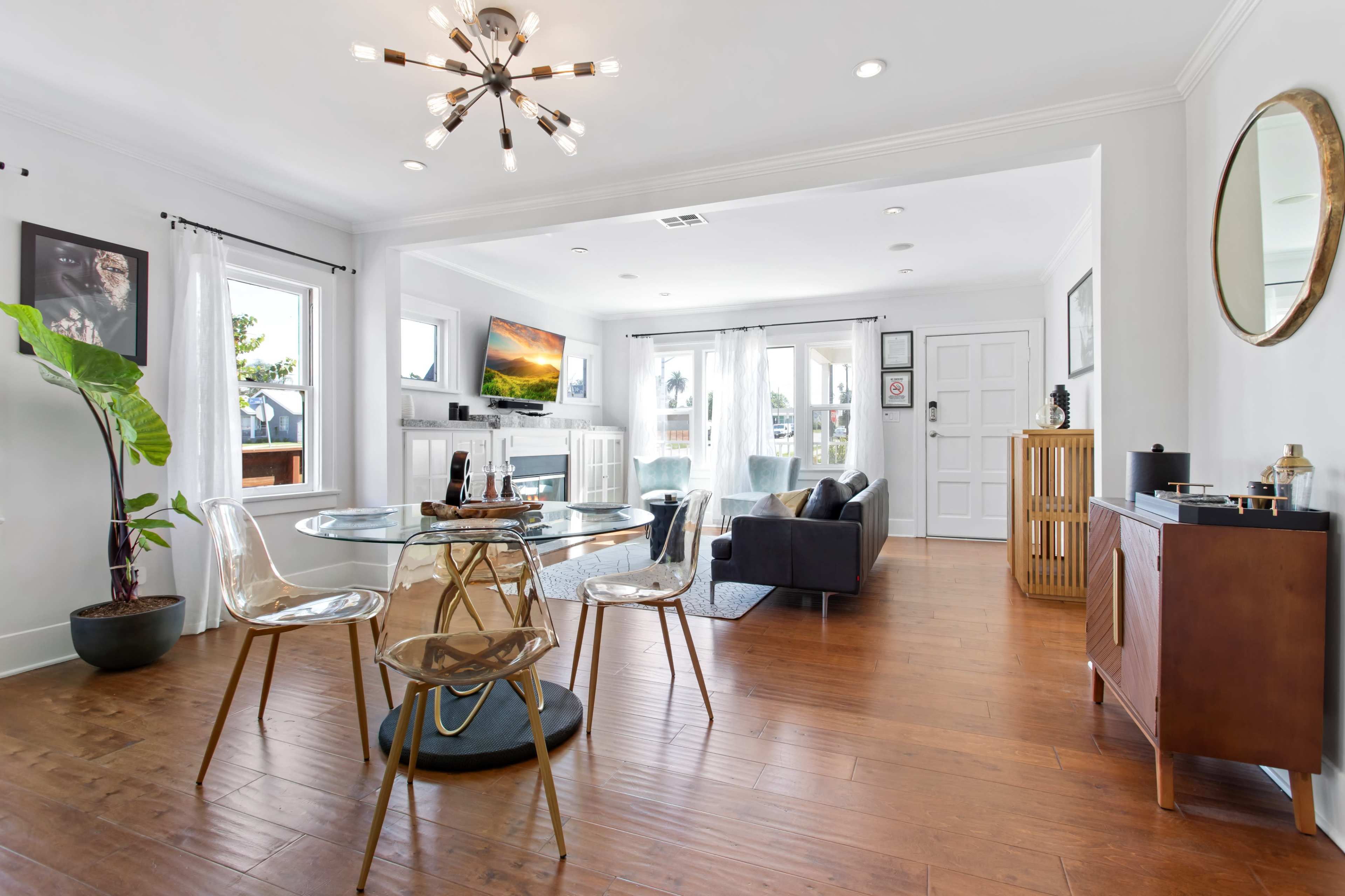 A modern living space features a circular glass dining table, clear acrylic chairs, and a cozy seating area with a large window and artwork on the walls.