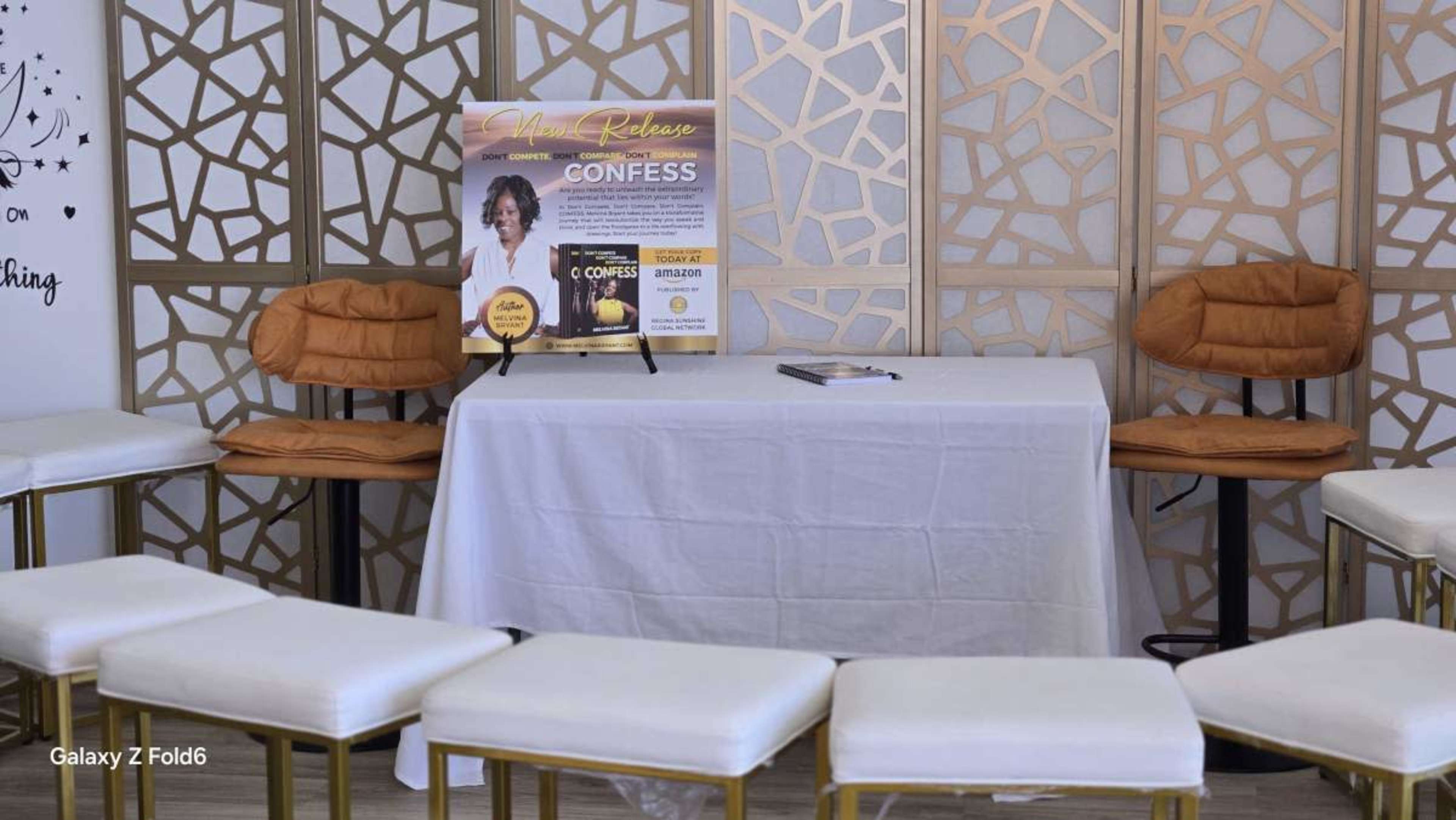 A table covered with a white cloth is set up in front of two chairs, with a promotional sign for an event titled "Confess" displayed on it, surrounded by circular seating.