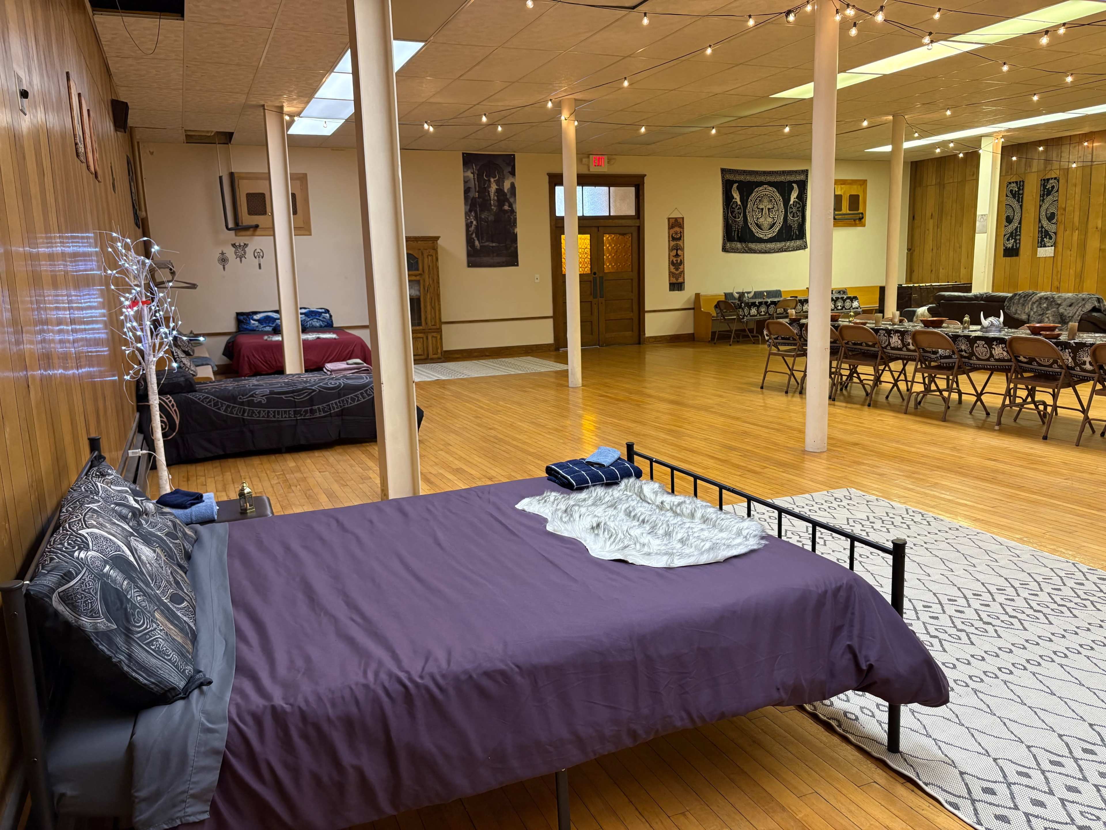 The image shows a spacious room featuring a bed with a purple blanket, surrounded by wooden flooring, various seating arrangements, and decorative items on the walls.