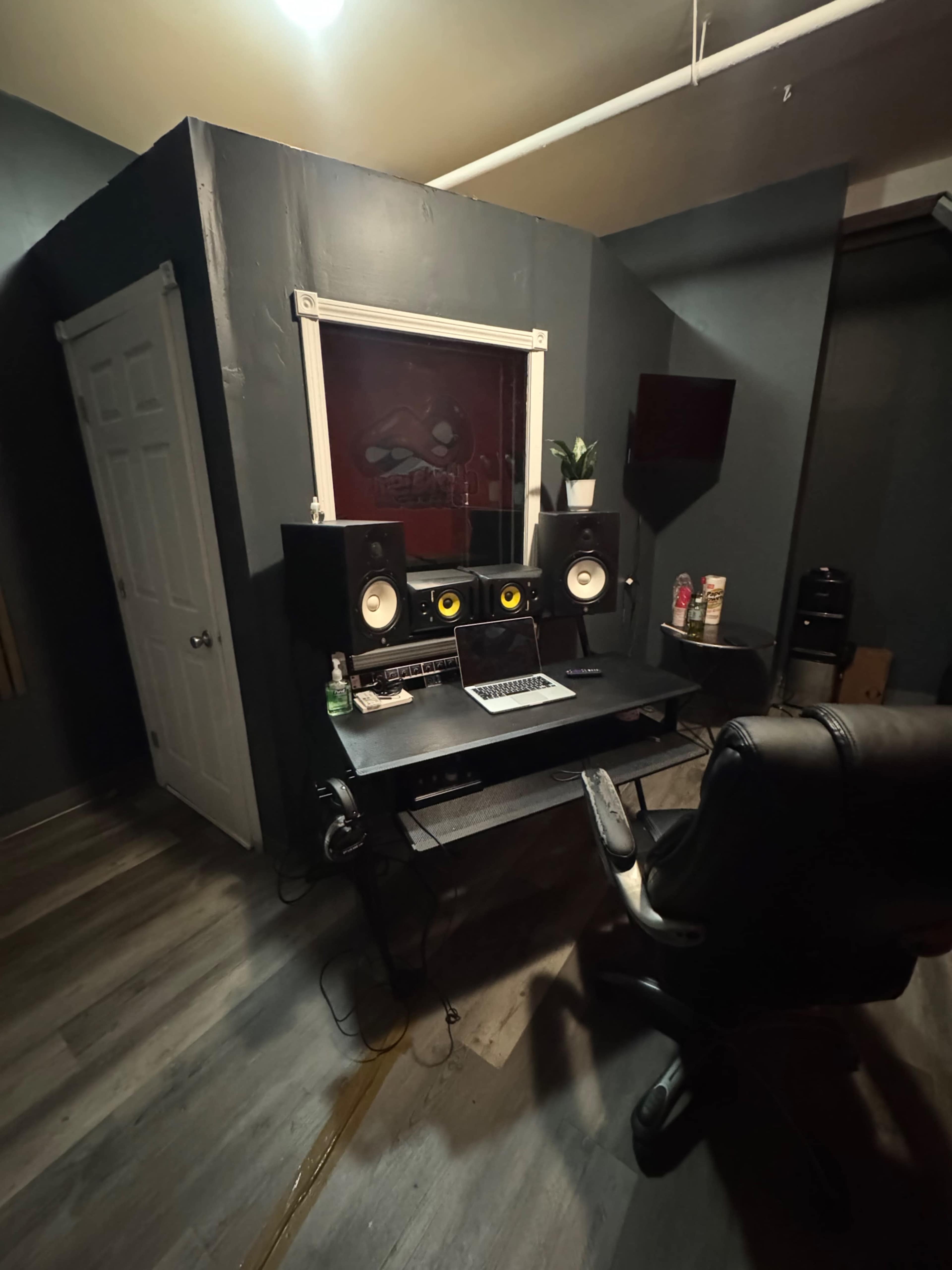 The image shows a music studio with a desk containing a computer and speakers, flanked by potted plants and a wall-mounted TV.