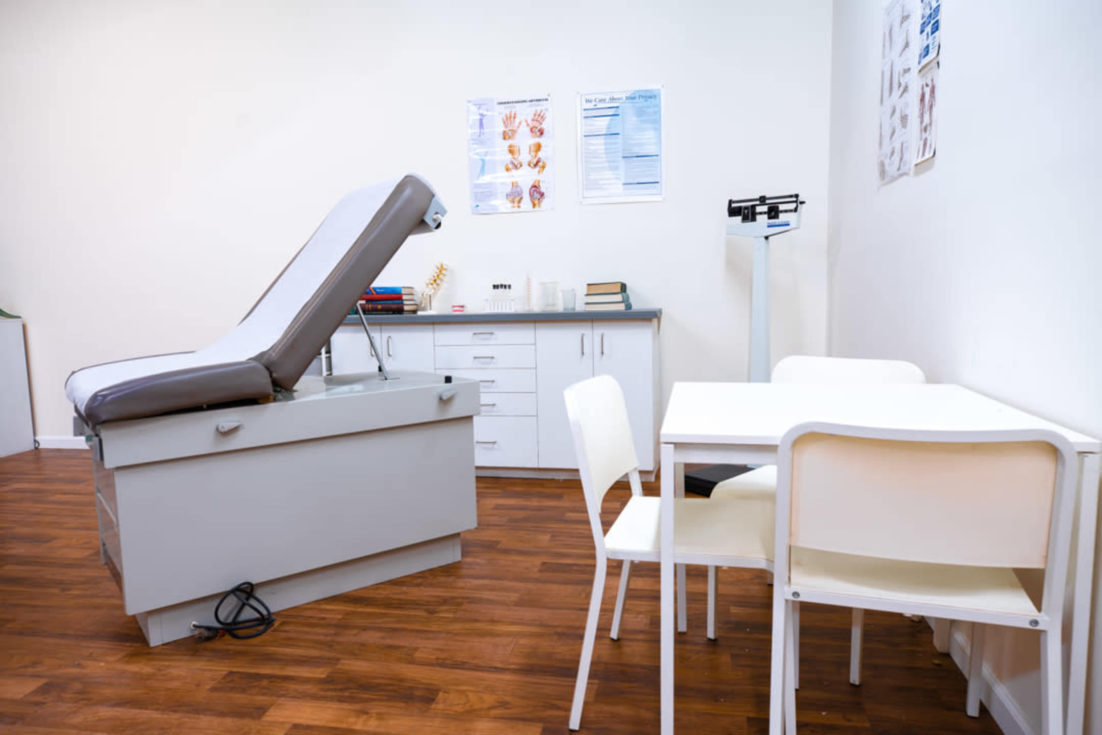 The image shows a medical examination room featuring an examination table, a storage cabinet, and a small table with chairs.