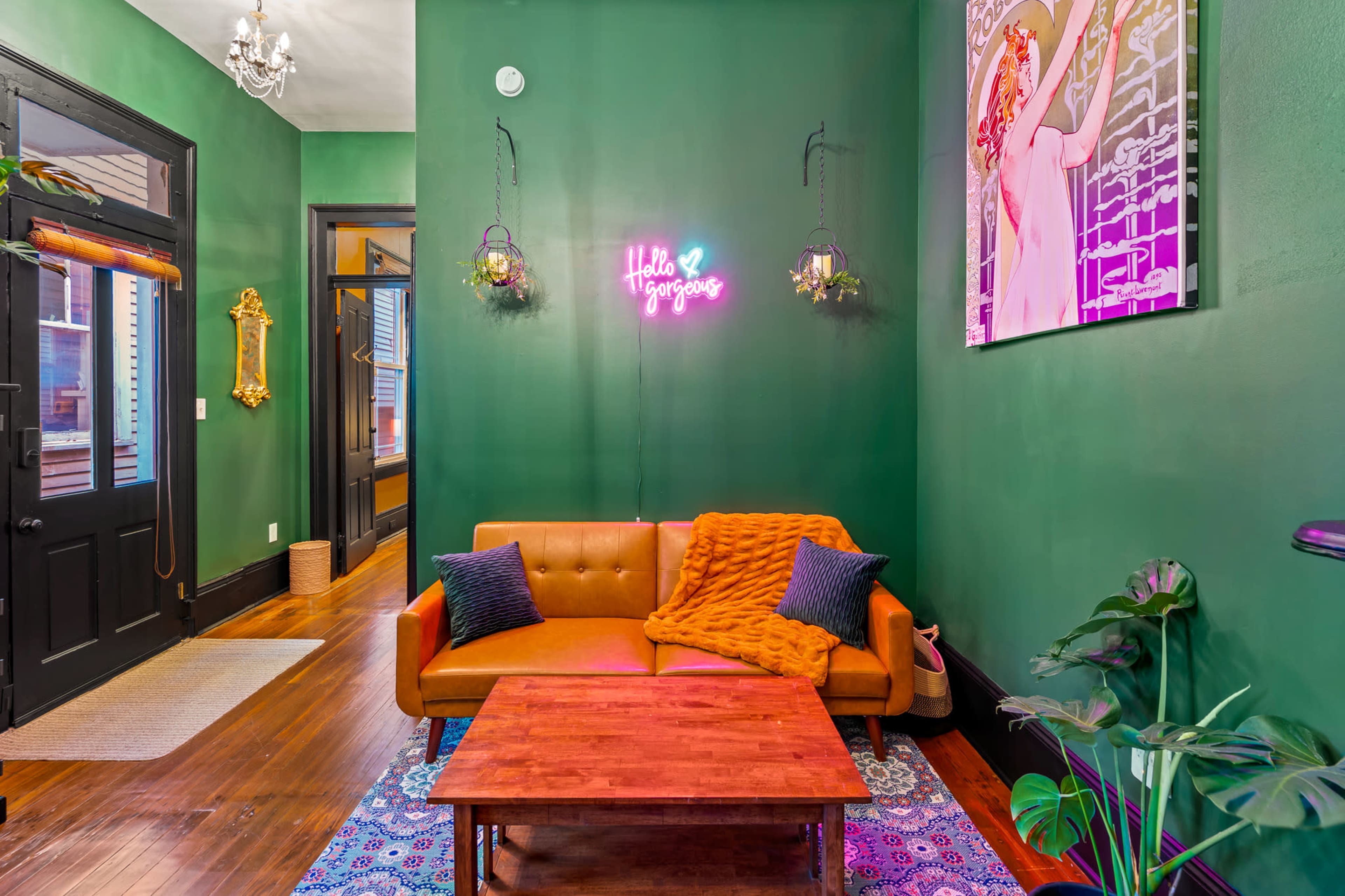 A vibrant room features a brown sofa with a colorful throw, a wooden coffee table, and green walls adorned with plants and a neon sign.