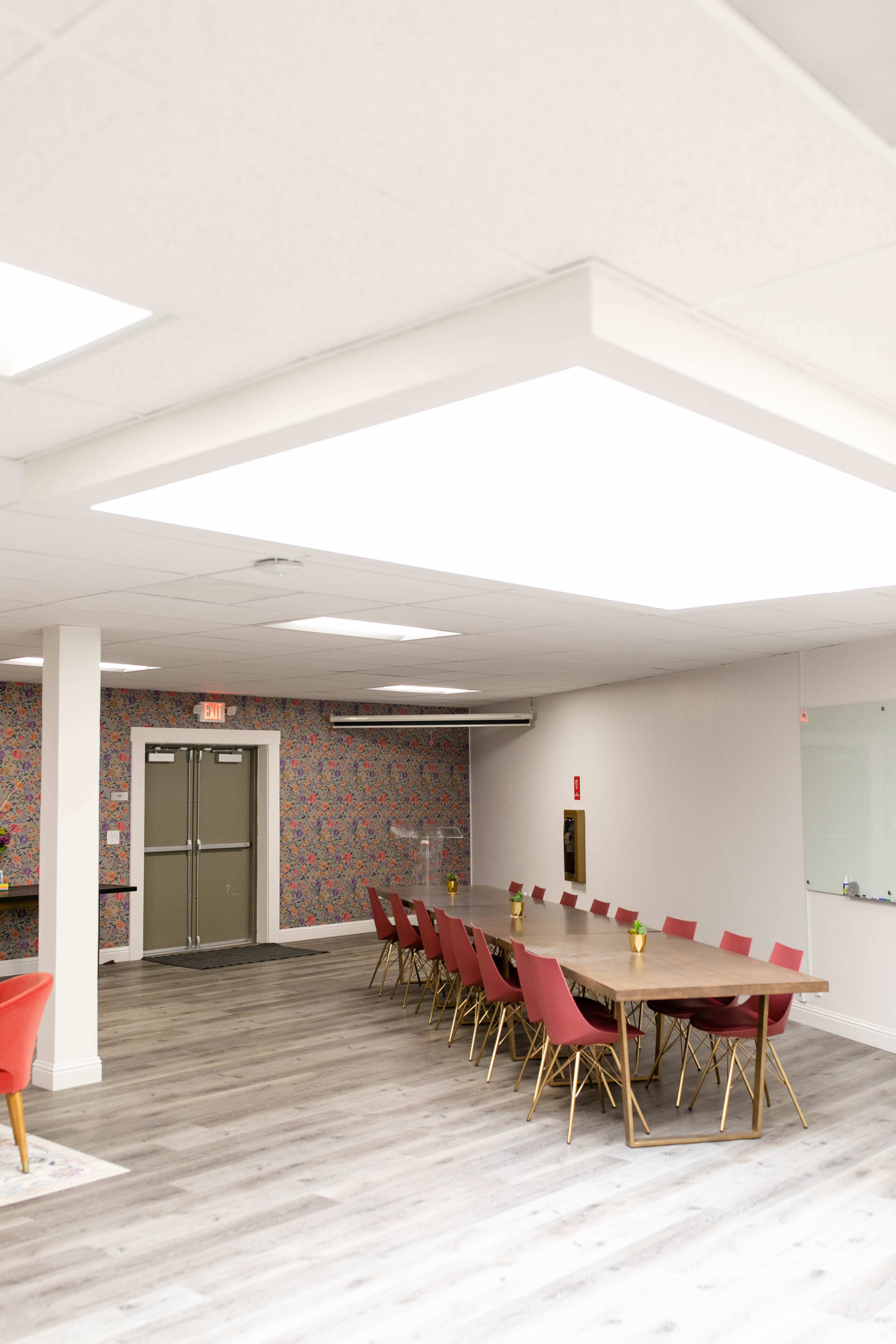 A spacious meeting room features a long wooden table surrounded by red chairs, set against a patterned floral wallpaper.