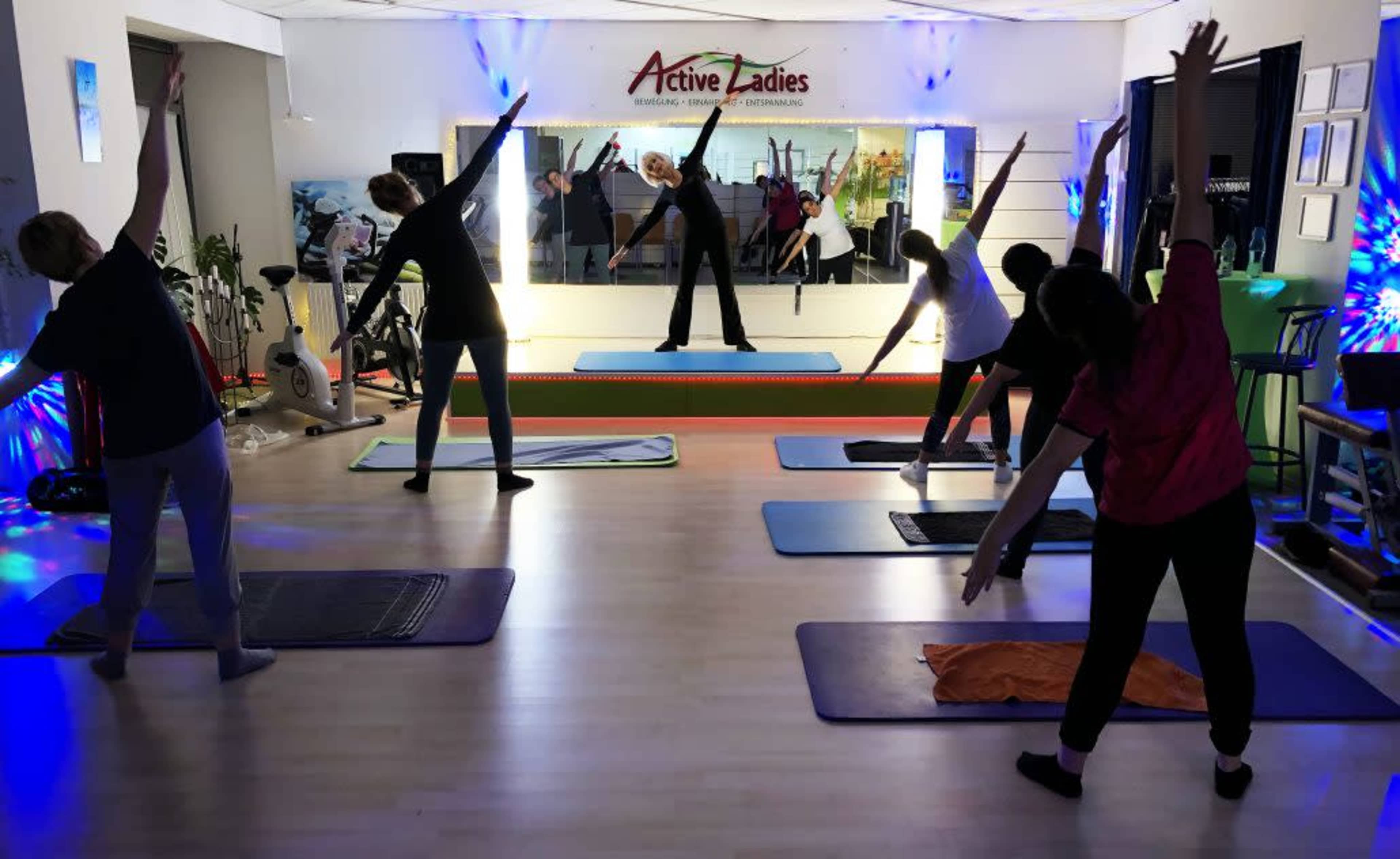 A group of women are participating in a fitness class, stretching with their arms raised, while an instructor leads in front of a mirror in a brightly lit studio.
