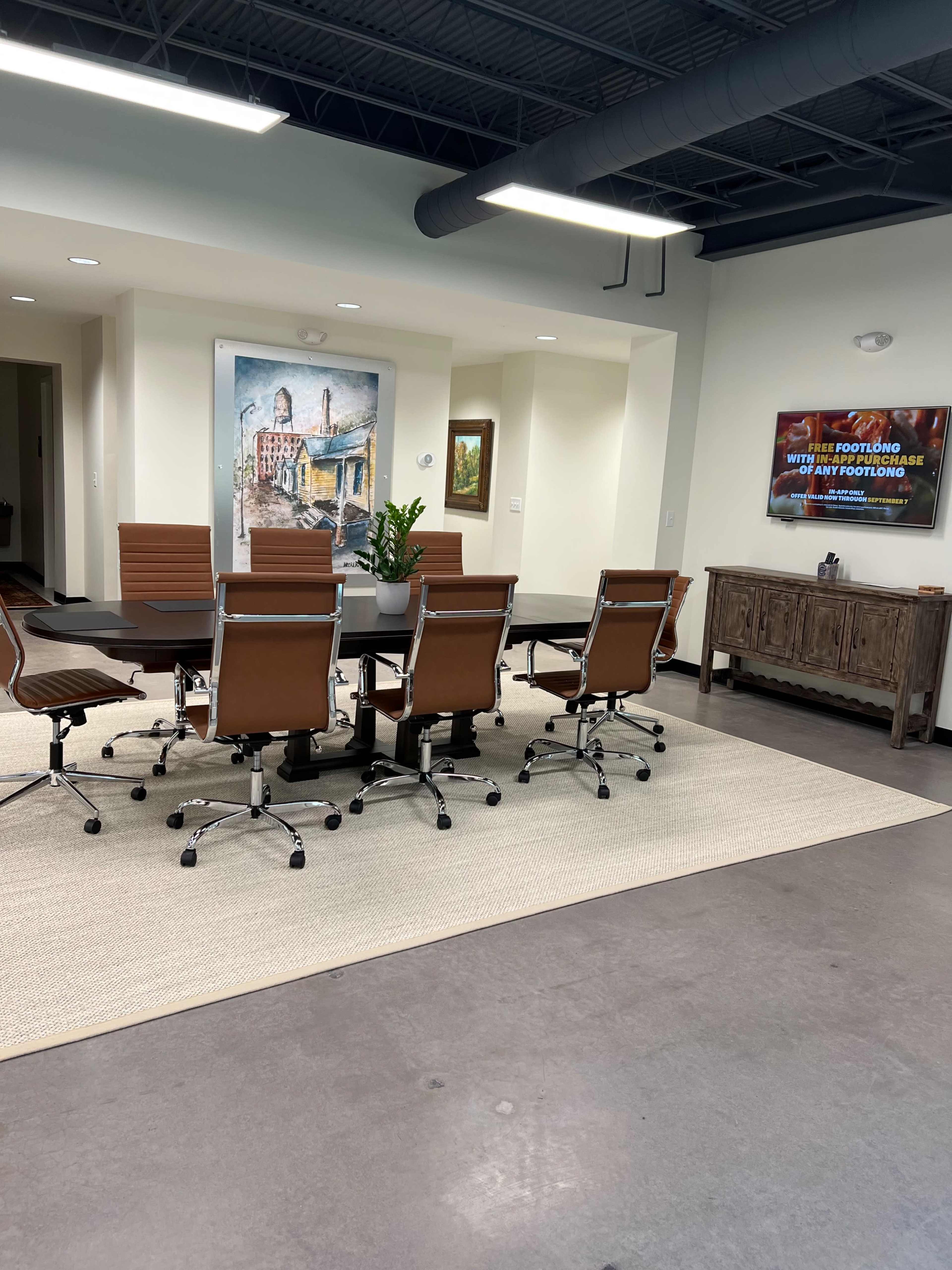 A conference room features a large table surrounded by brown leather chairs, with artwork and a plant enhancing the decor.