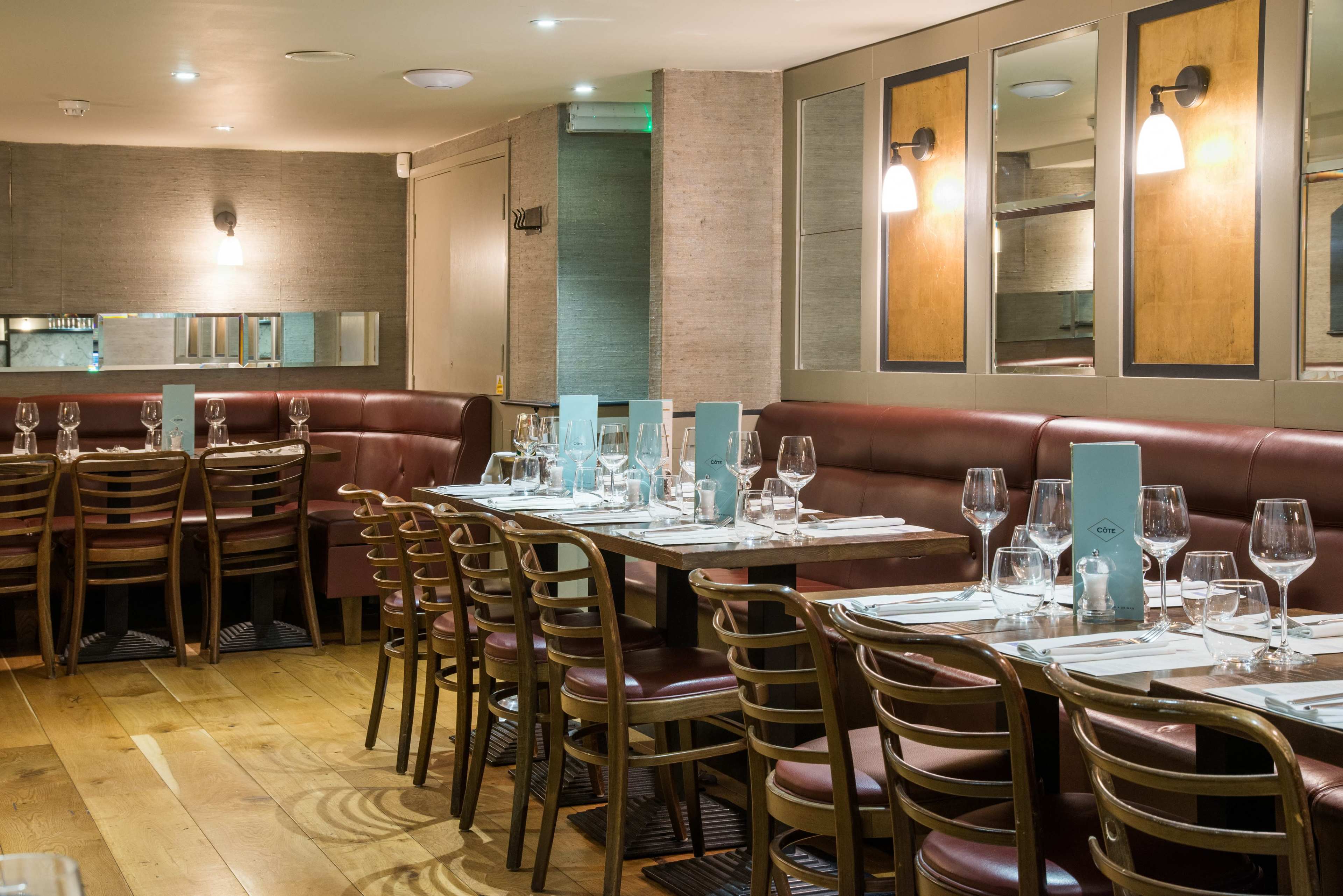 The image shows a restaurant interior featuring several tables set with glasses and utensils, surrounded by red leather seating and wooden flooring.