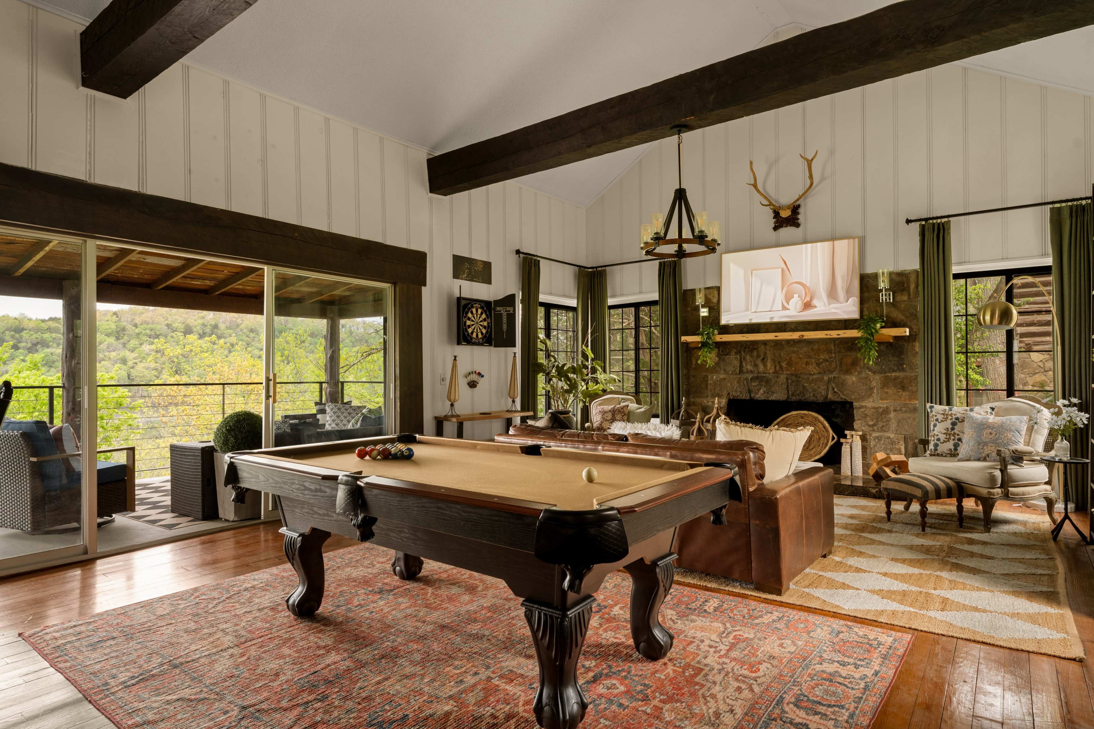 The image shows a cozy living room with a billiard table, a stone fireplace, and large windows overlooking a green landscape.