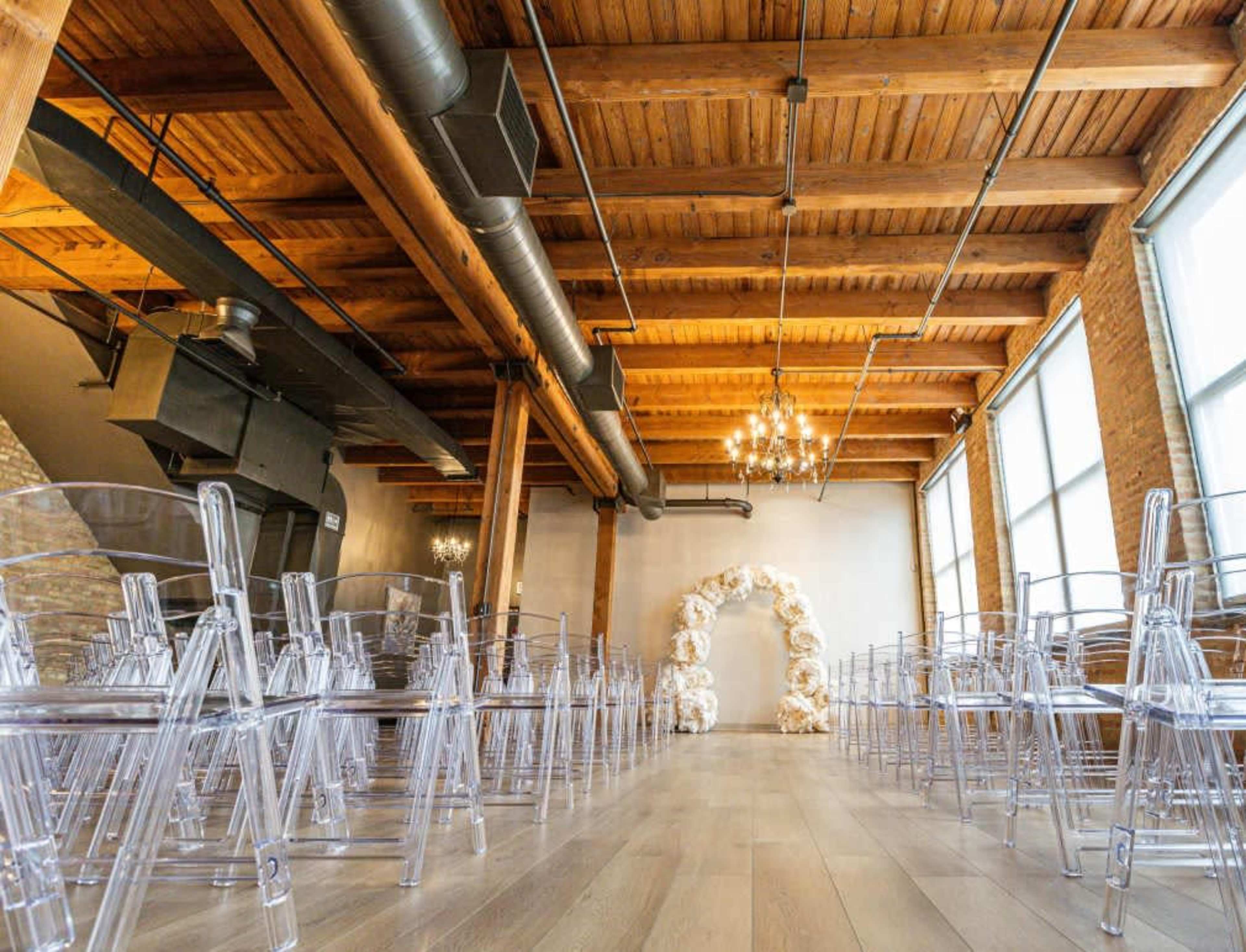 The image shows an indoor event space with wooden beams, large windows, and a decorative balloon arch at one end, with rows of clear plastic chairs arranged facing it.