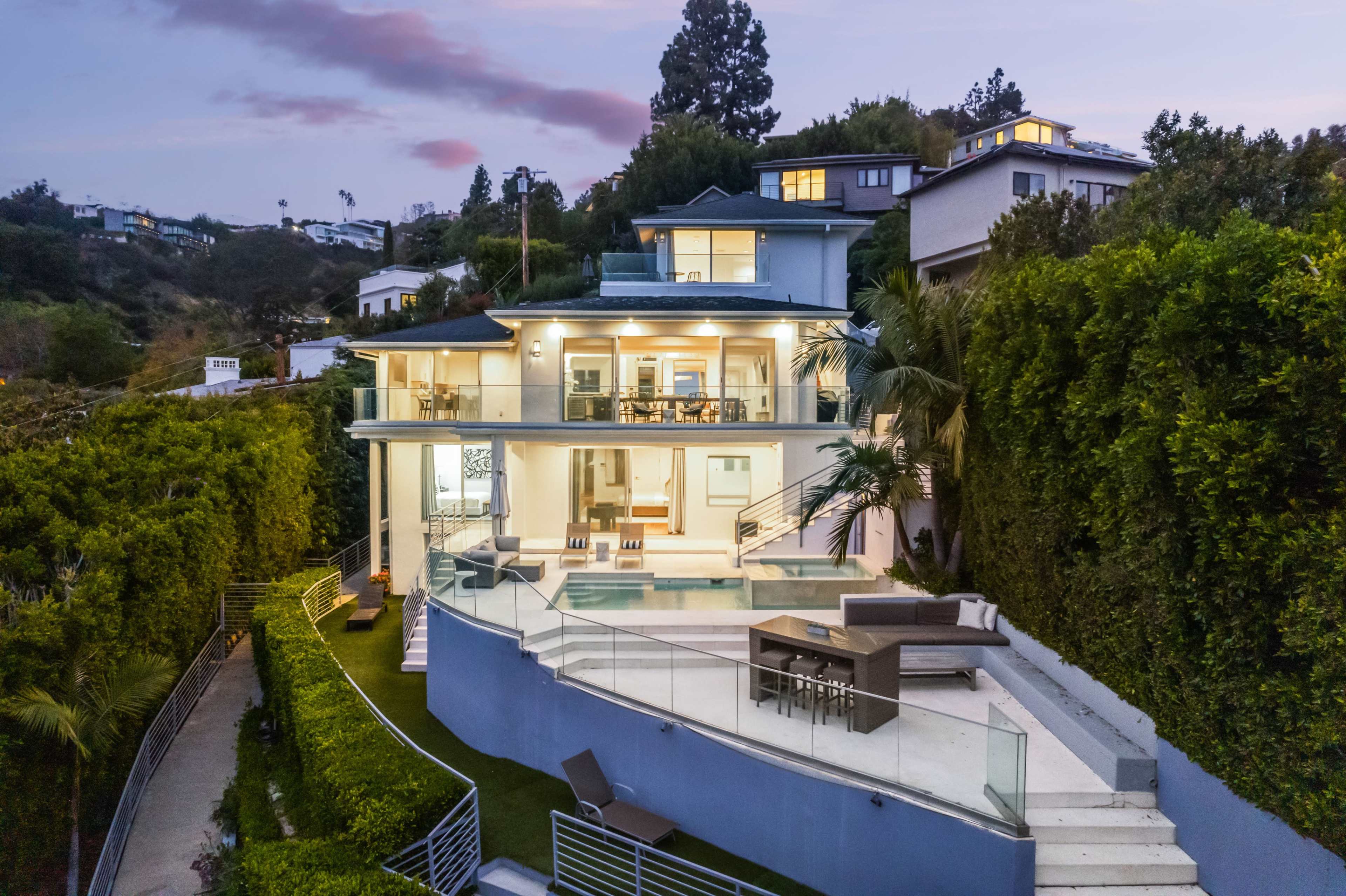 A modern multi-level home features a large glass-walled living area, a swimming pool, and lush landscaping in the surrounding hillside.