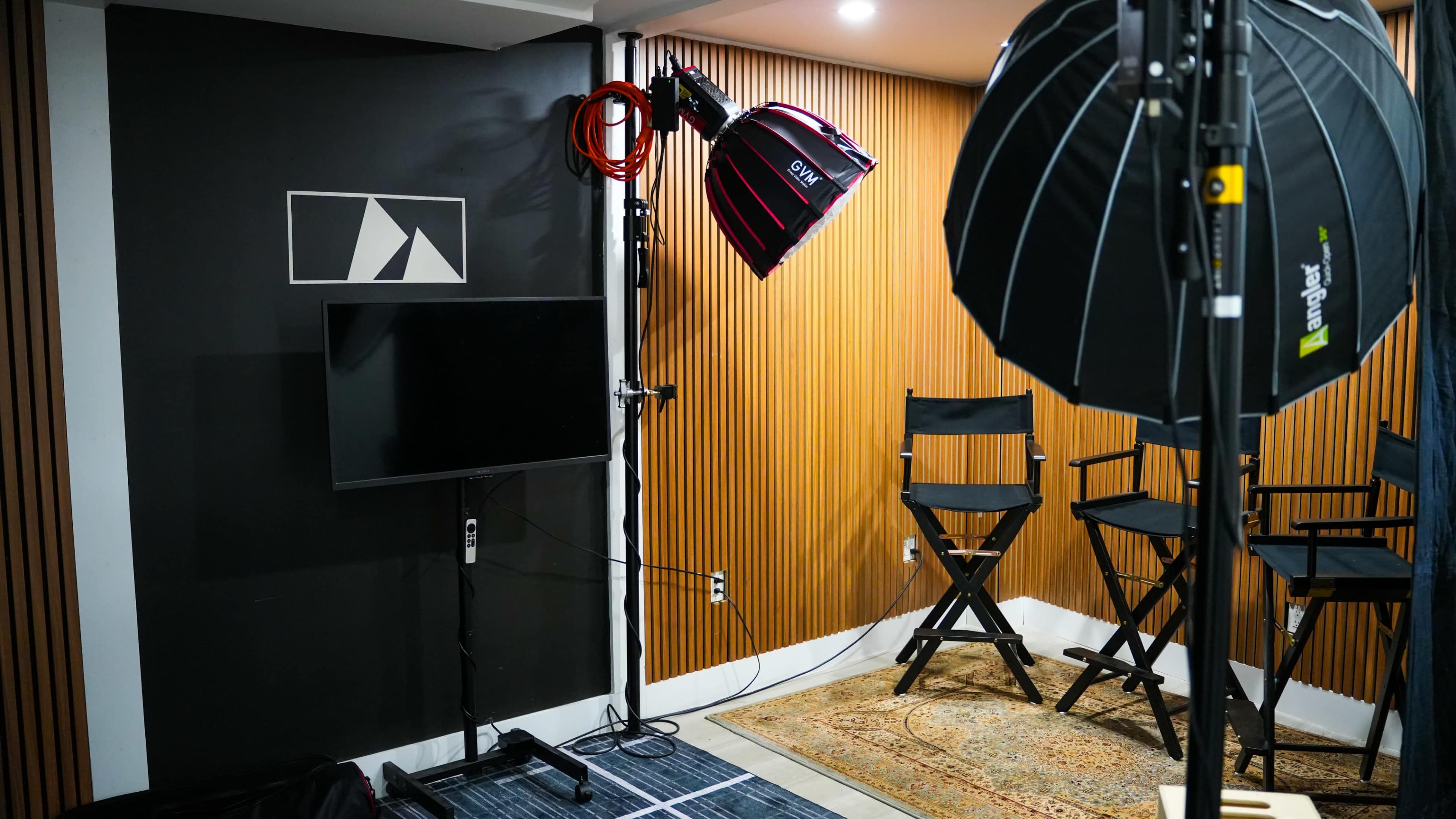 The image shows a small studio setup with a television screen, two director's chairs, and professional lighting equipment against a wooden panel backdrop.