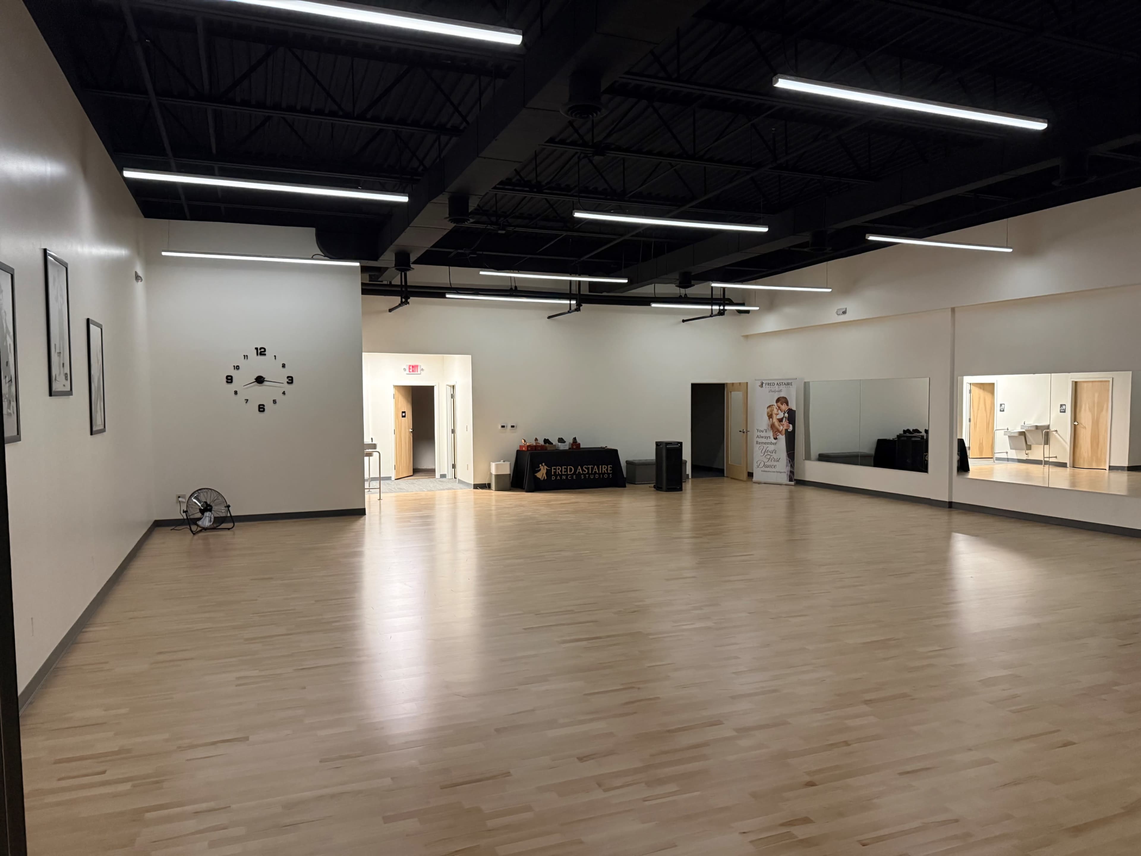 The image shows a spacious dance studio with wooden flooring, mirrors on one wall, and a reception area at the back.