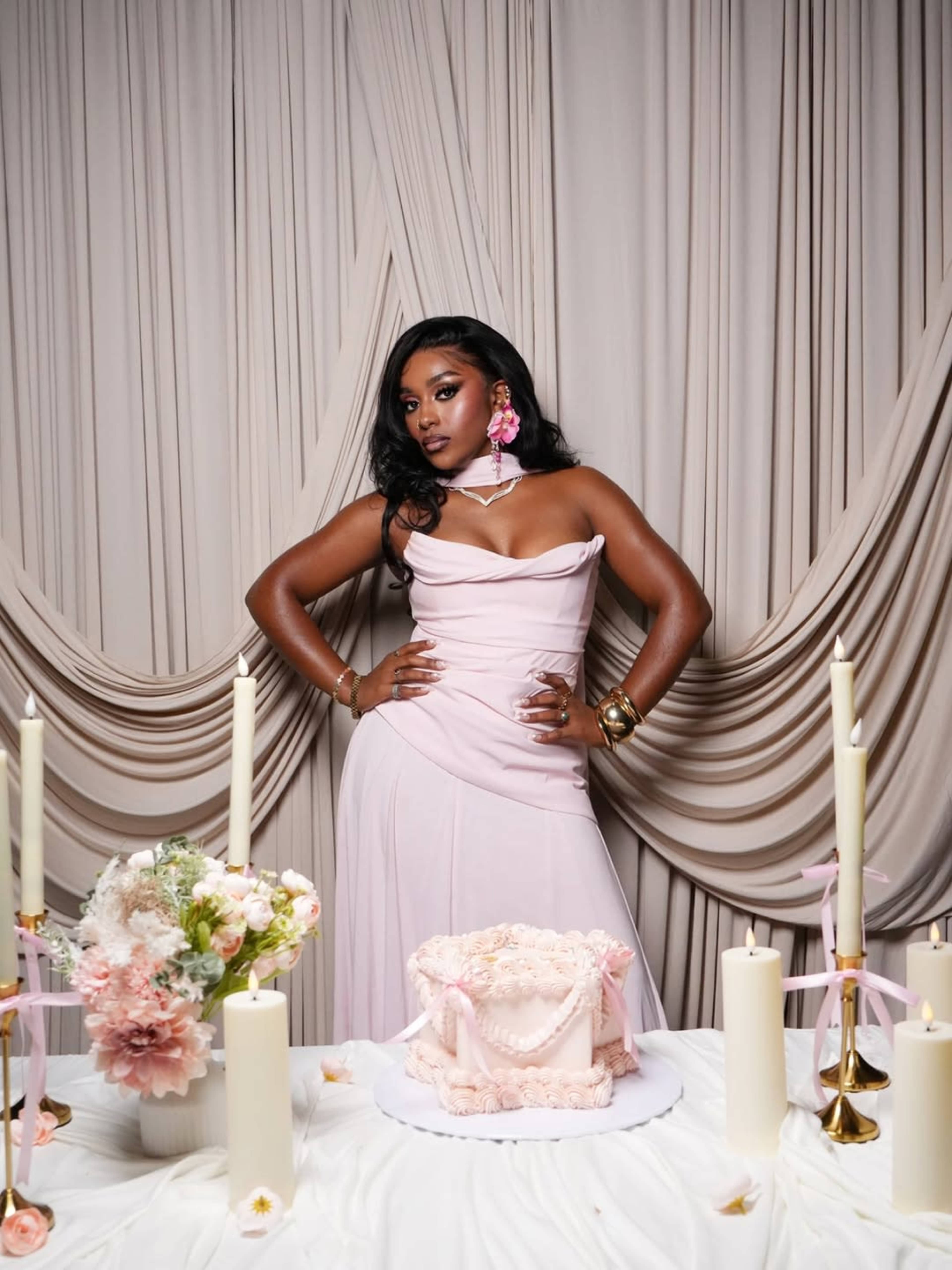 A woman in a light pink dress stands confidently in front of a table adorned with a decorated cake, candles, and floral arrangements against a backdrop of draped fabric.