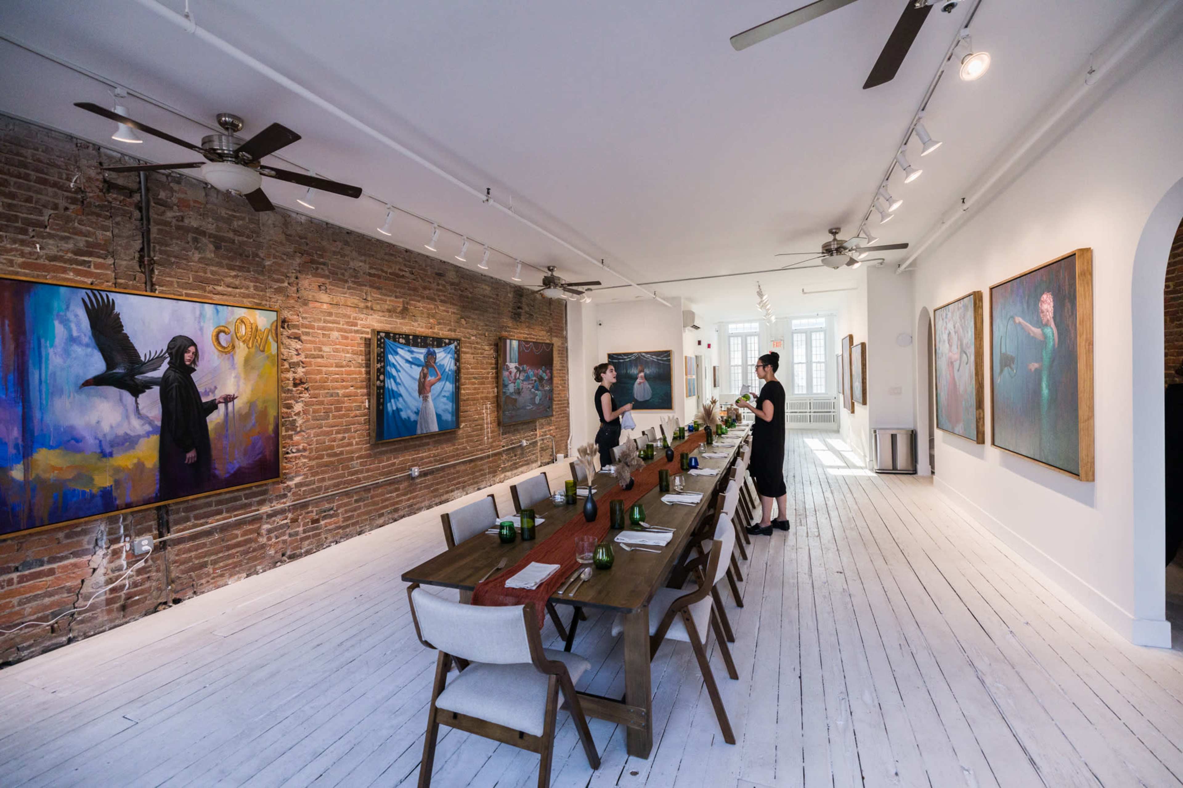 A long wooden dining table is set with greenery in a gallery featuring large paintings on exposed brick walls.