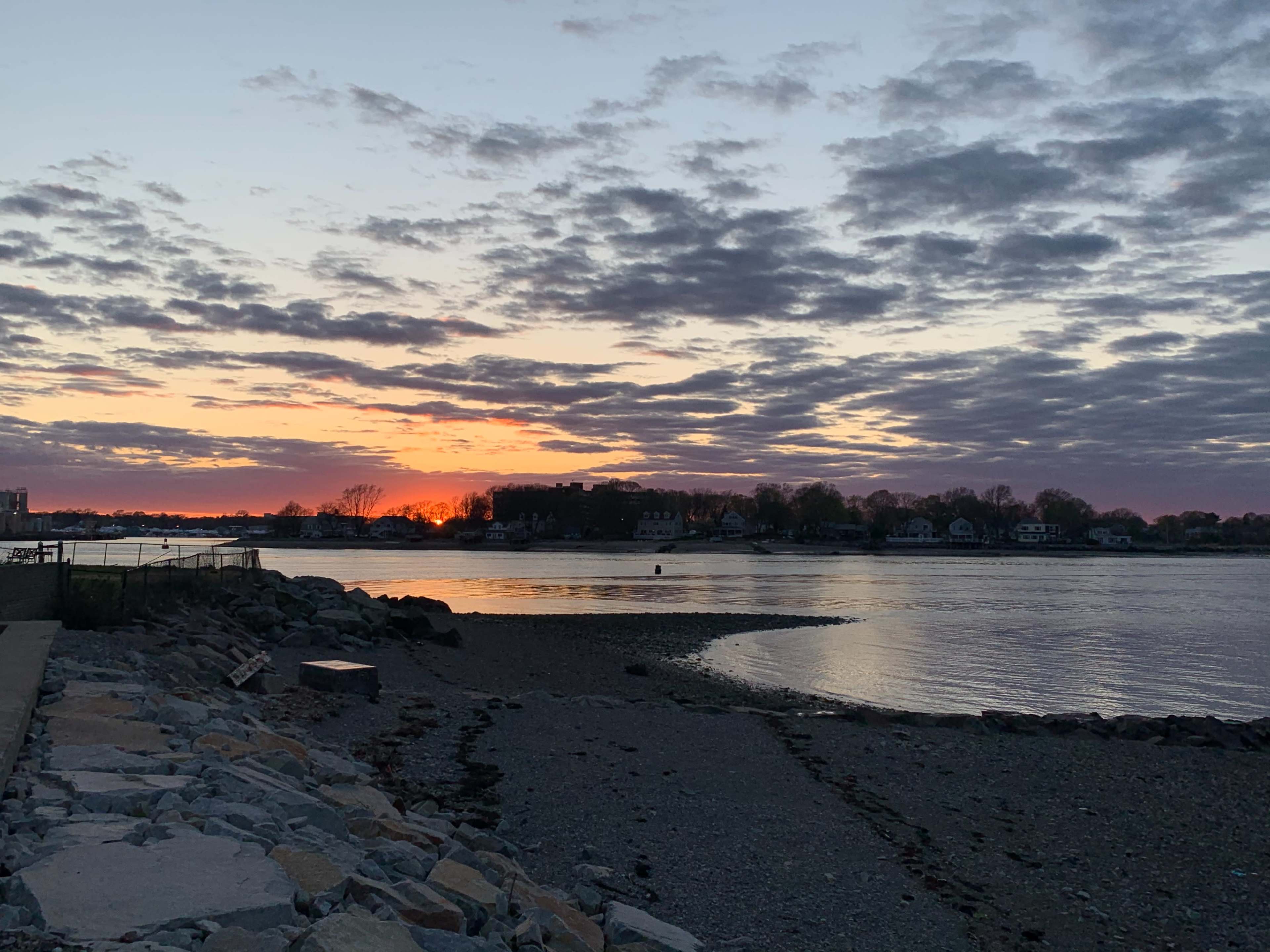 The image shows a sunset over a calm body of water, with clouds in the sky and a rocky shore in the foreground.