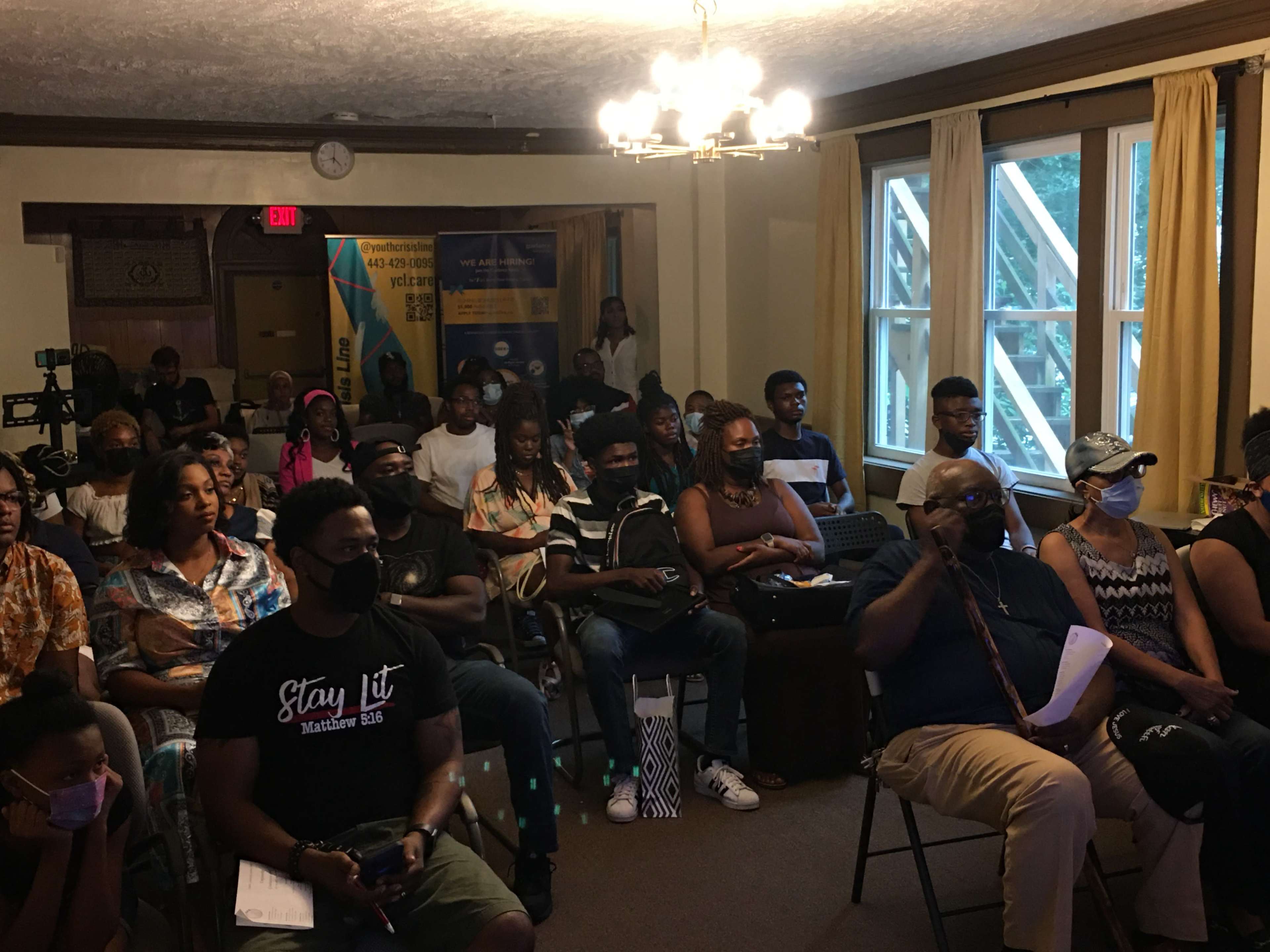 A diverse group of people is seated in a room for an event, with some wearing masks and various attendees engaged in discussions or activities.