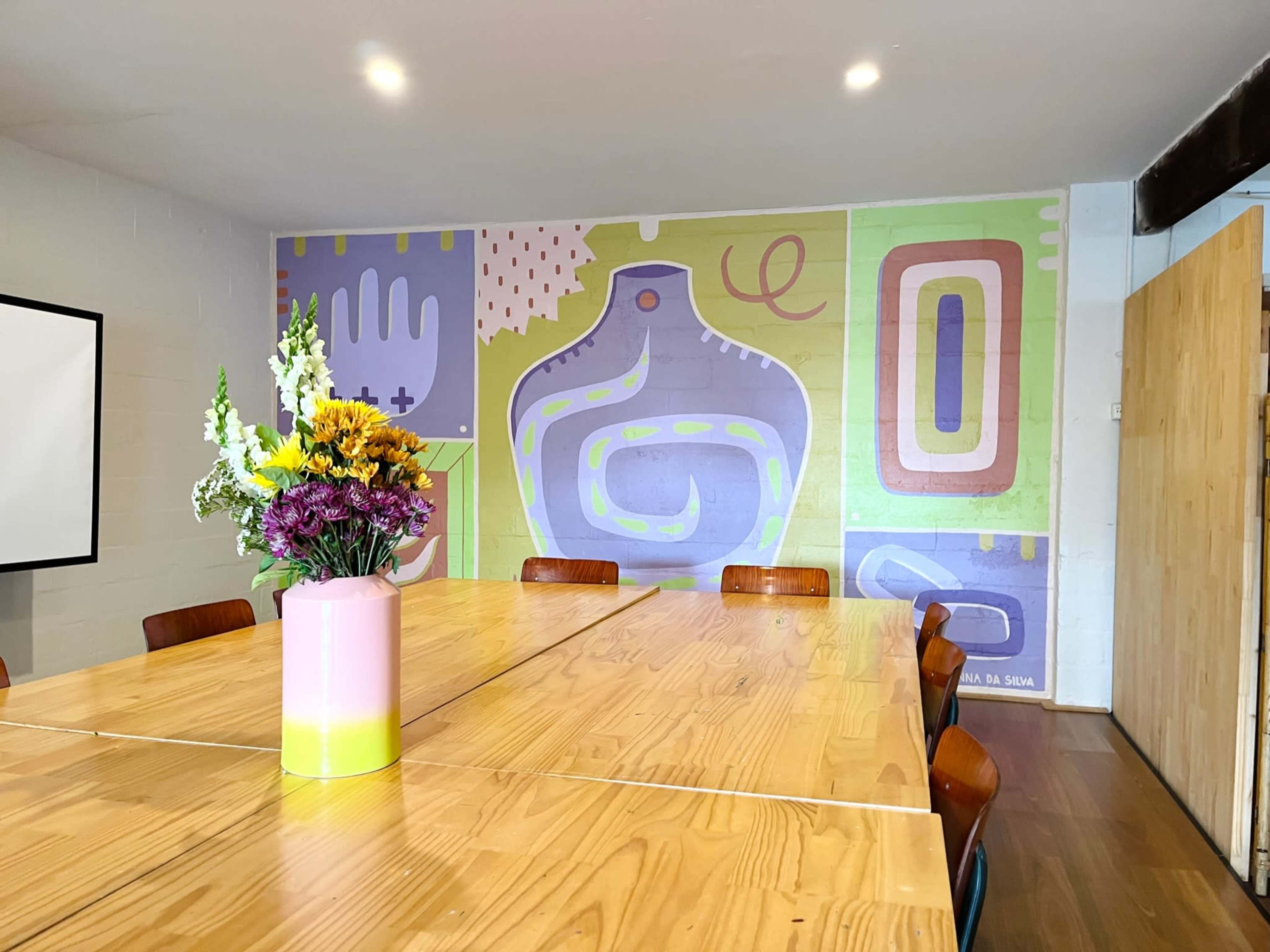 A long wooden table with several chairs is arranged in a brightly lit room featuring a colorful mural on the wall and a vase of flowers in the center.