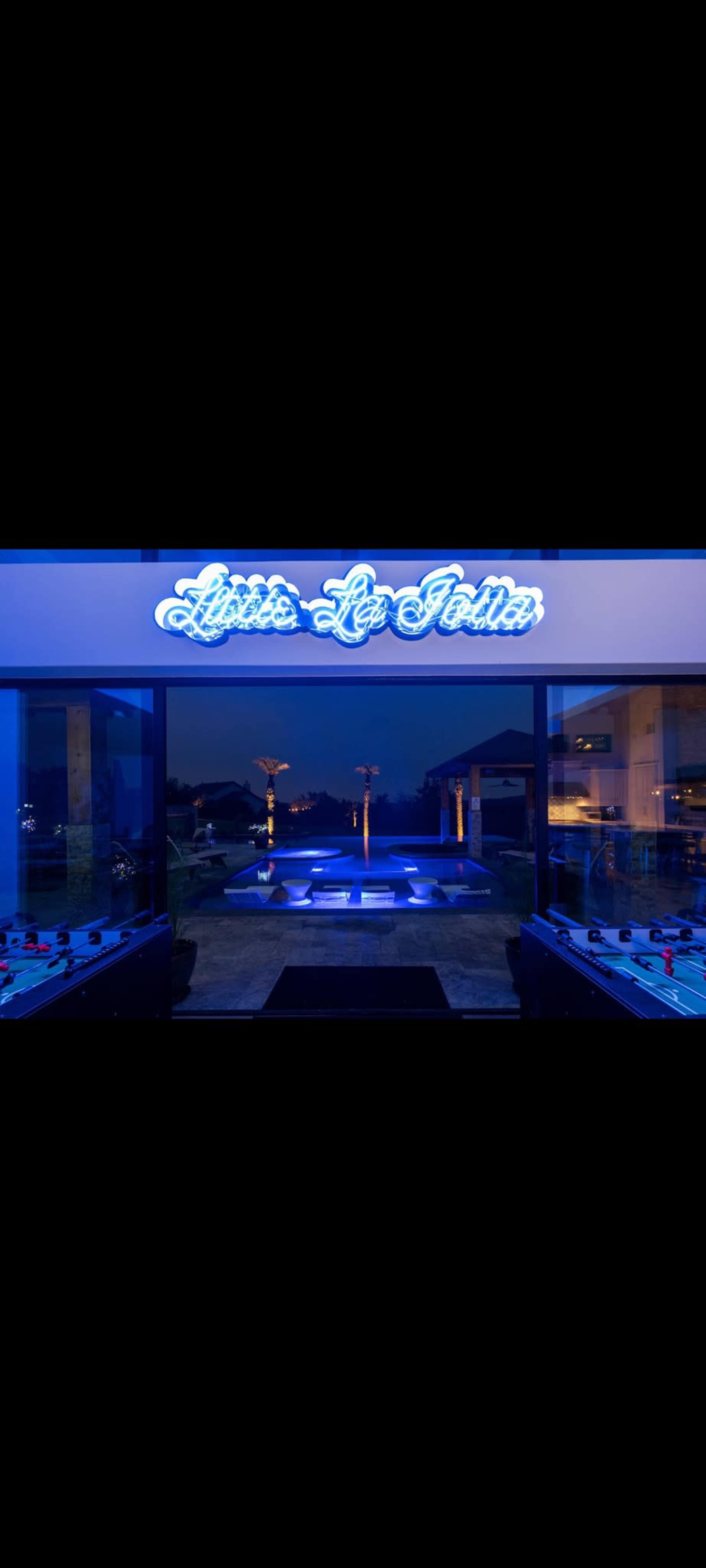 The image shows a modern entrance featuring a neon sign that reads "Lydia La Jolla" and a view of an illuminated outdoor area with a pool.