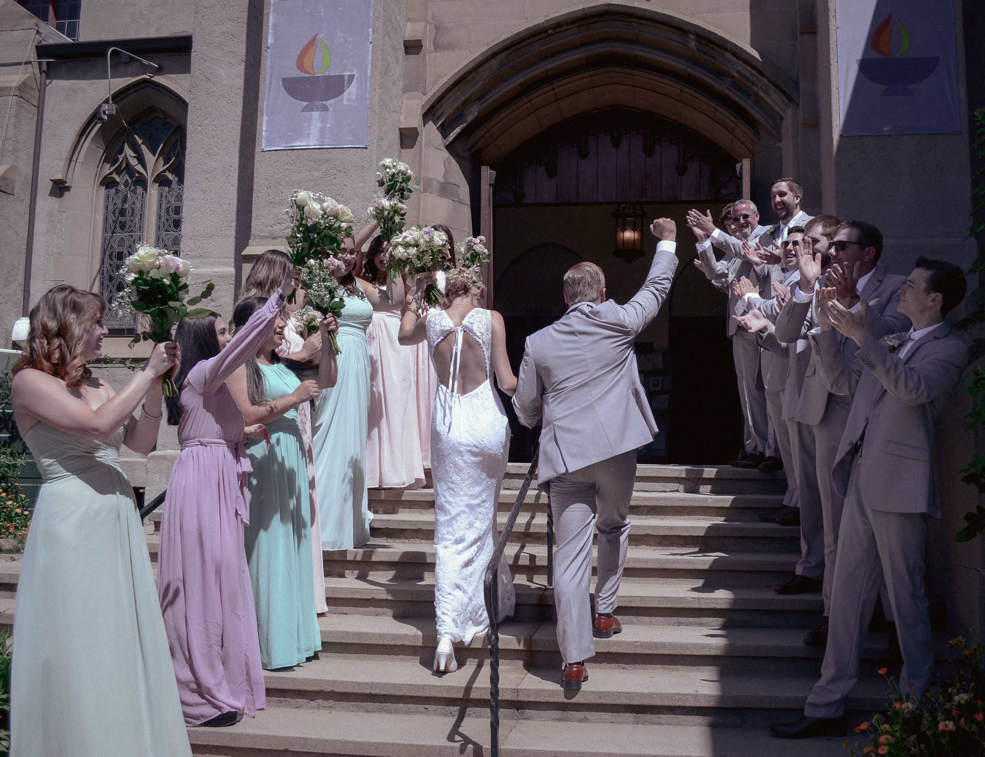 A wedding party stands on the steps of a church, with the bride and groom ascending while guests cheer and hold bouquets.
