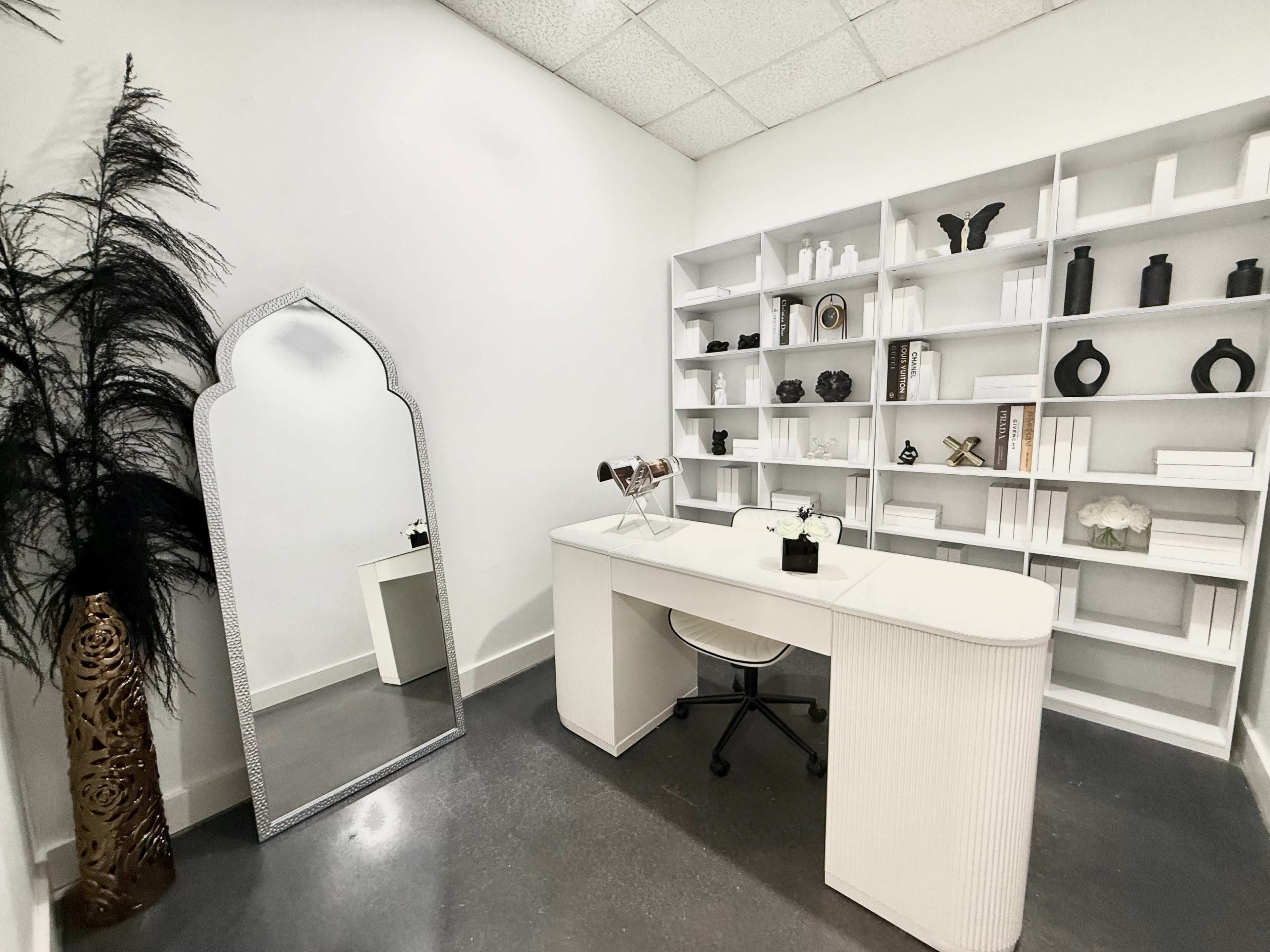 The image showcases a minimalist office space featuring a white desk and chair, a large decorative mirror, and neatly arranged shelves with various decorative items.