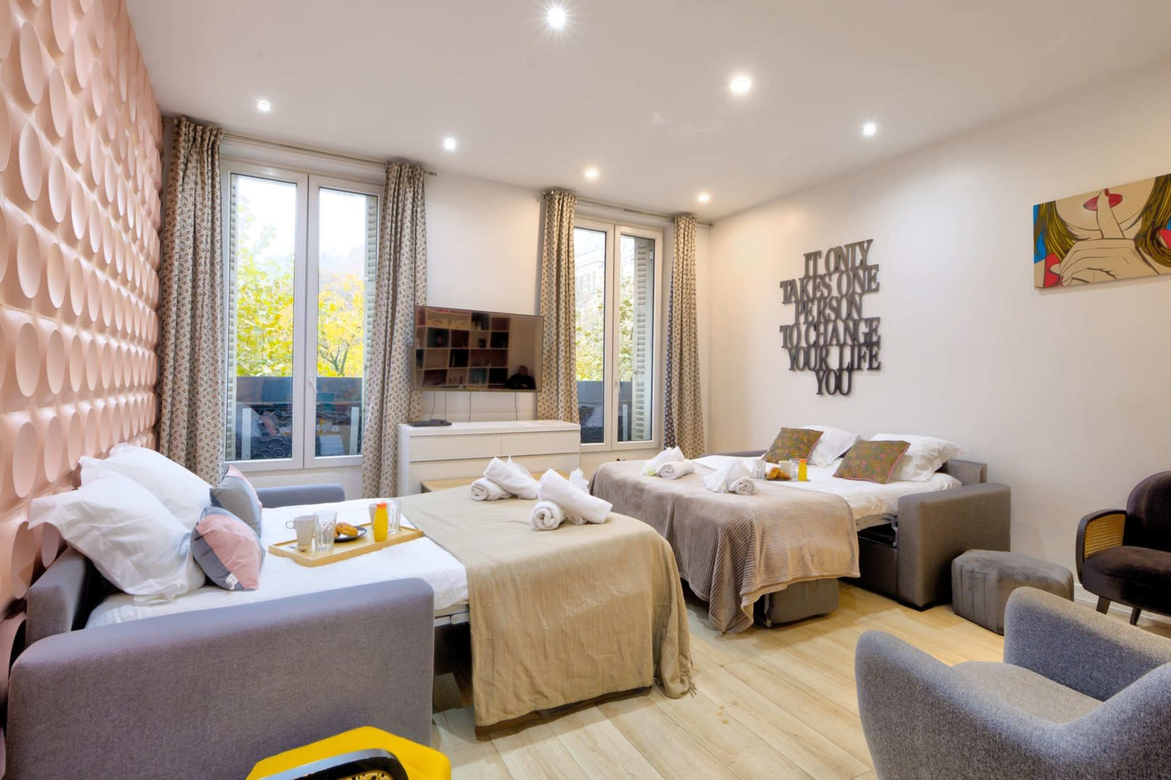 The room features two beds with light bedding, a television on the wall, and large windows allowing natural light to enter.