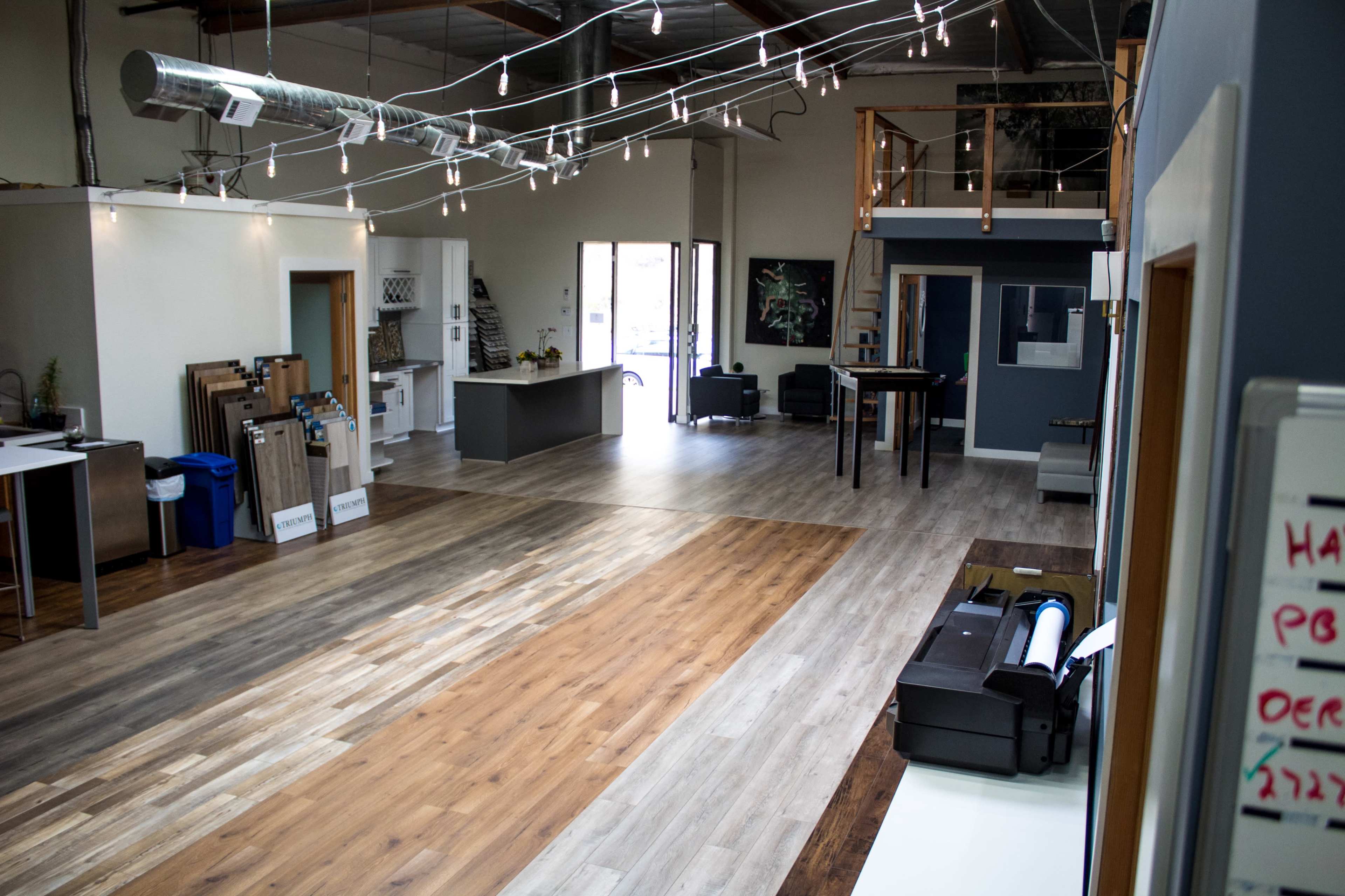 A modern interior space with various flooring samples displayed, a kitchen area, and a seating area lit by decorative string lights.