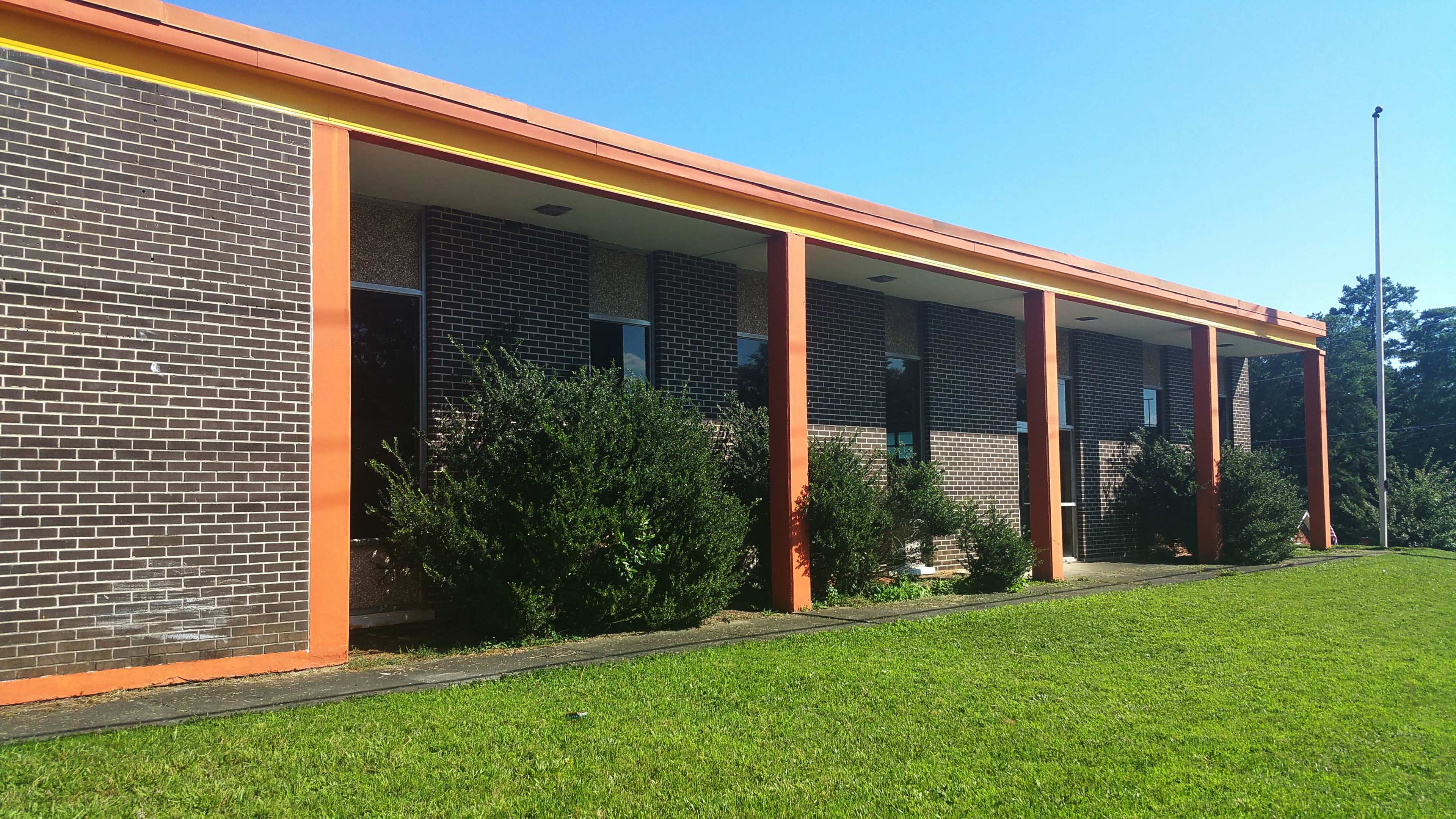 A single-story building with a brick facade and orange trim, set against a clear blue sky and surrounded by green grass and bushes.