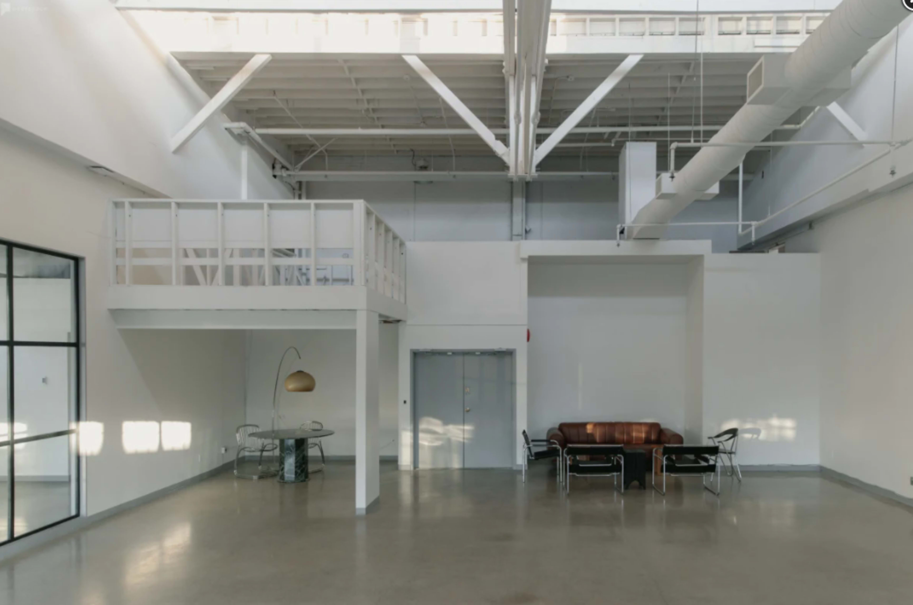 An expansive, modern interior space with a lofted area, a brown leather sofa, a dining table, and significant natural light entering through large windows.