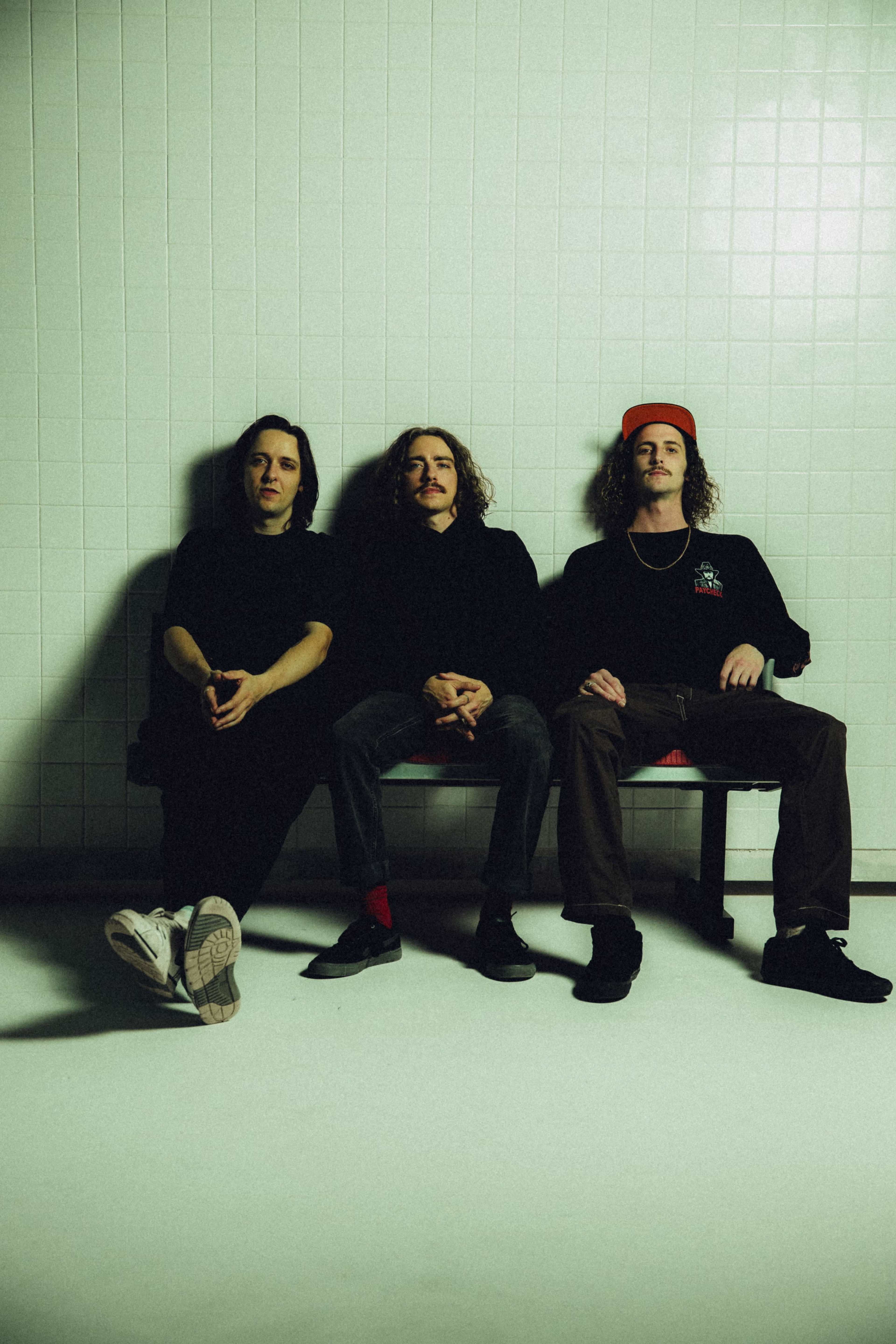 Three individuals sit together on a bench in a minimalist, tiled room.