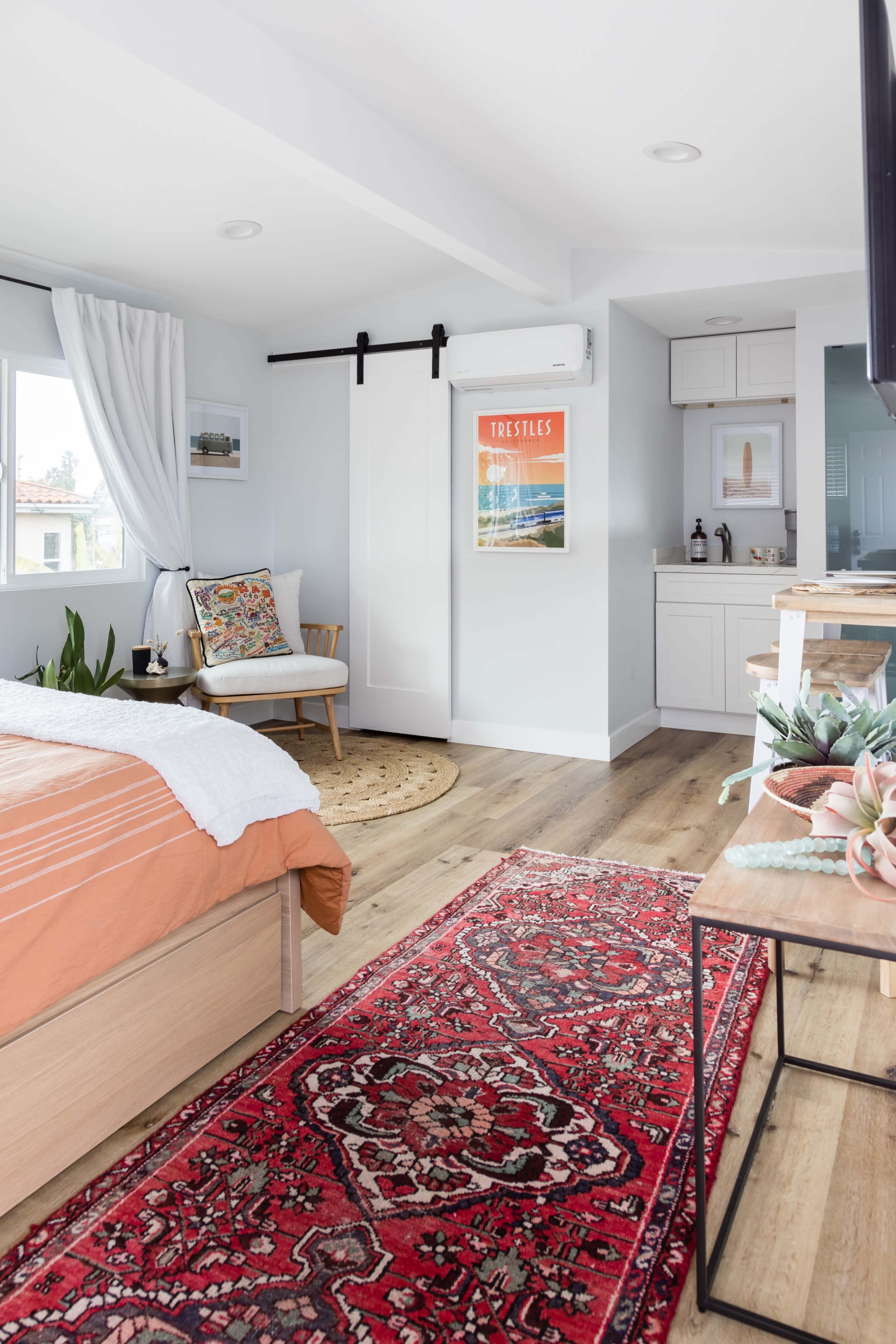The image shows a stylish bedroom featuring a bed with orange bedding, a decorative chair, a sliding barn door, and a red patterned rug on wooden flooring.
