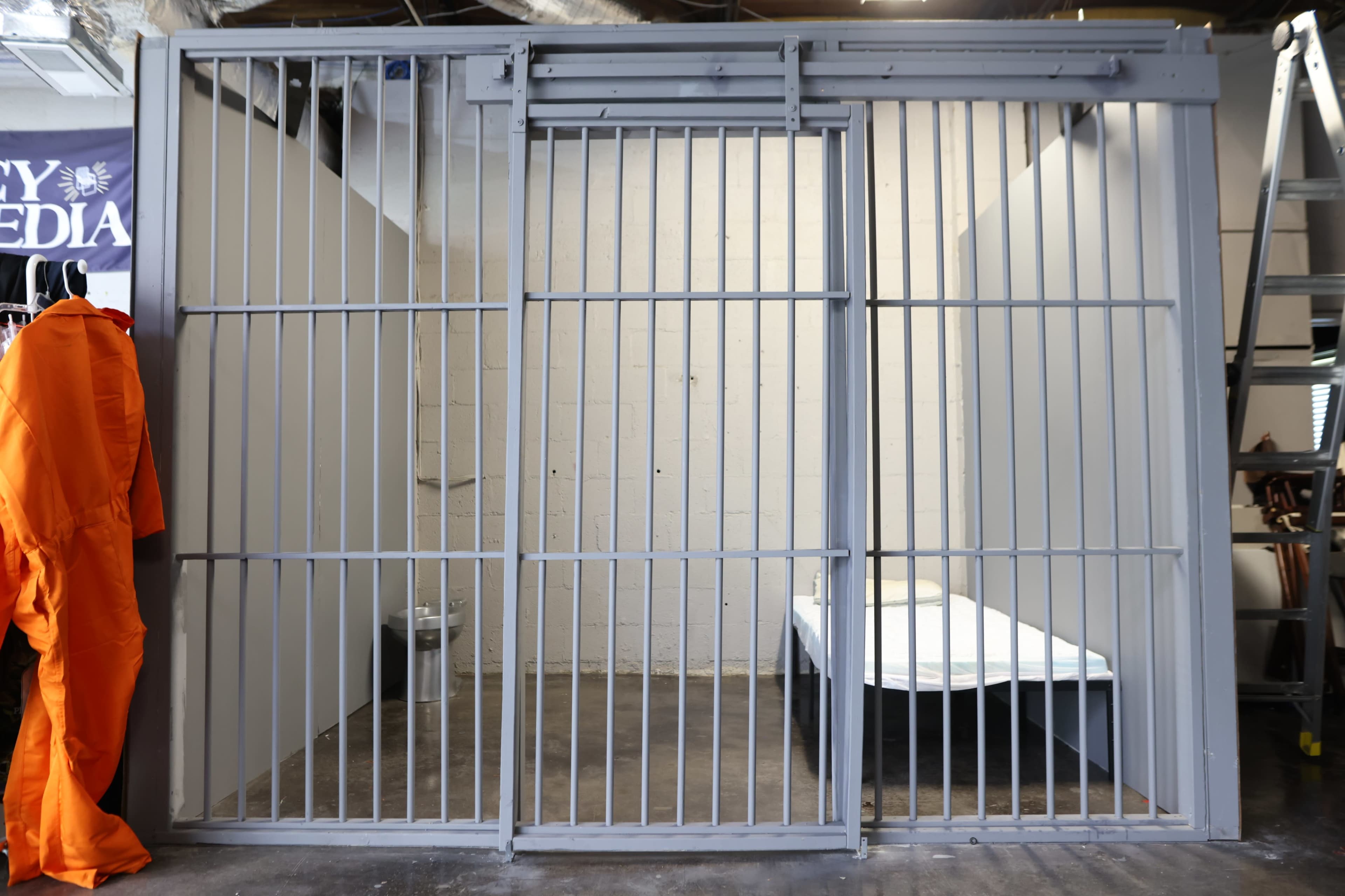 The image shows a jail cell with metal bars, a single bed inside, and an orange jumpsuit hanging on the side.