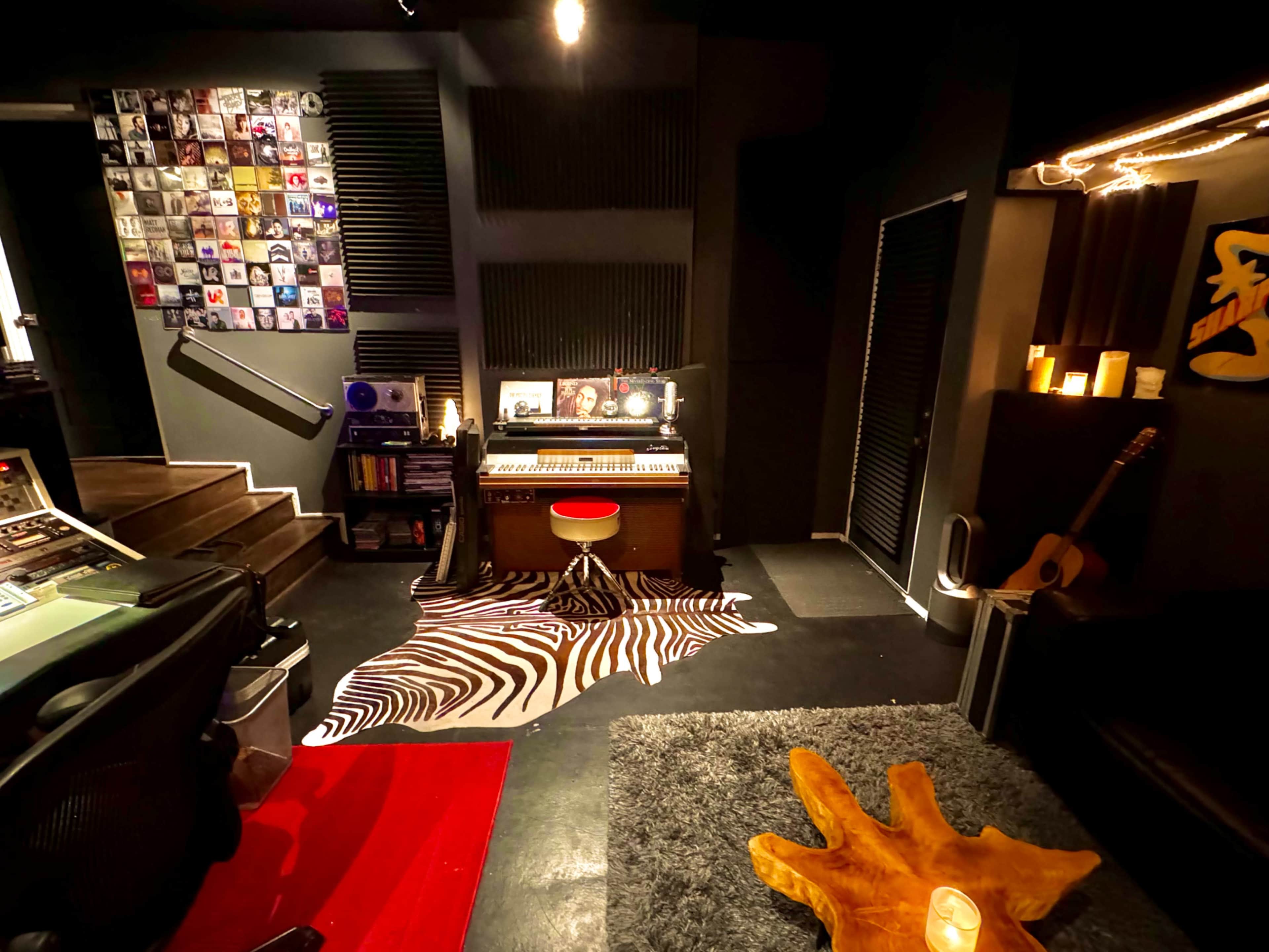 The image shows a music recording studio with a keyboard on a zebra-striped rug, a wall of vinyl records, and a cozy seating area.