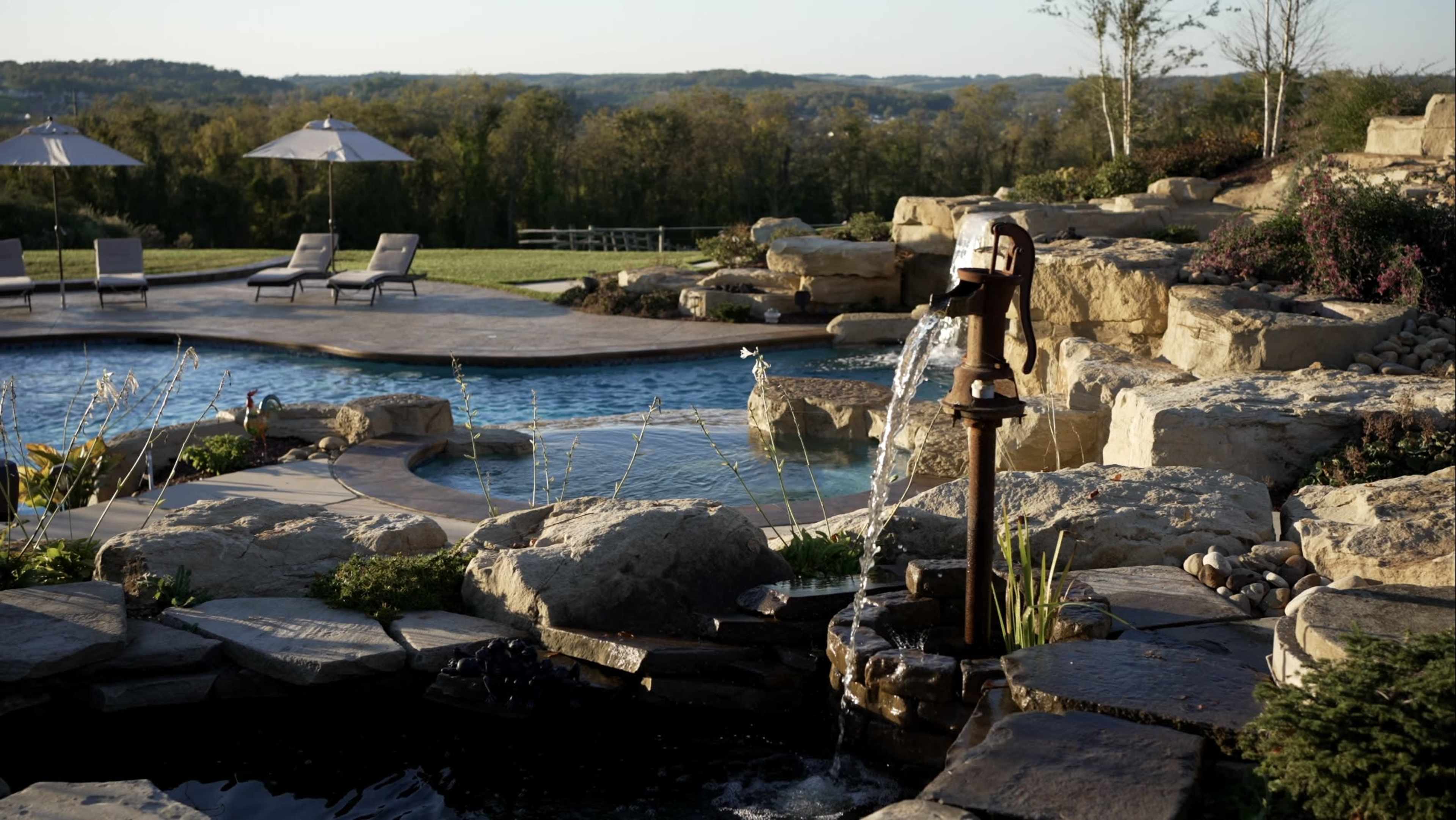 A stone-finished pool area features a water fountain, surrounded by landscaped rocks and lounge chairs, with a scenic backdrop of rolling hills.