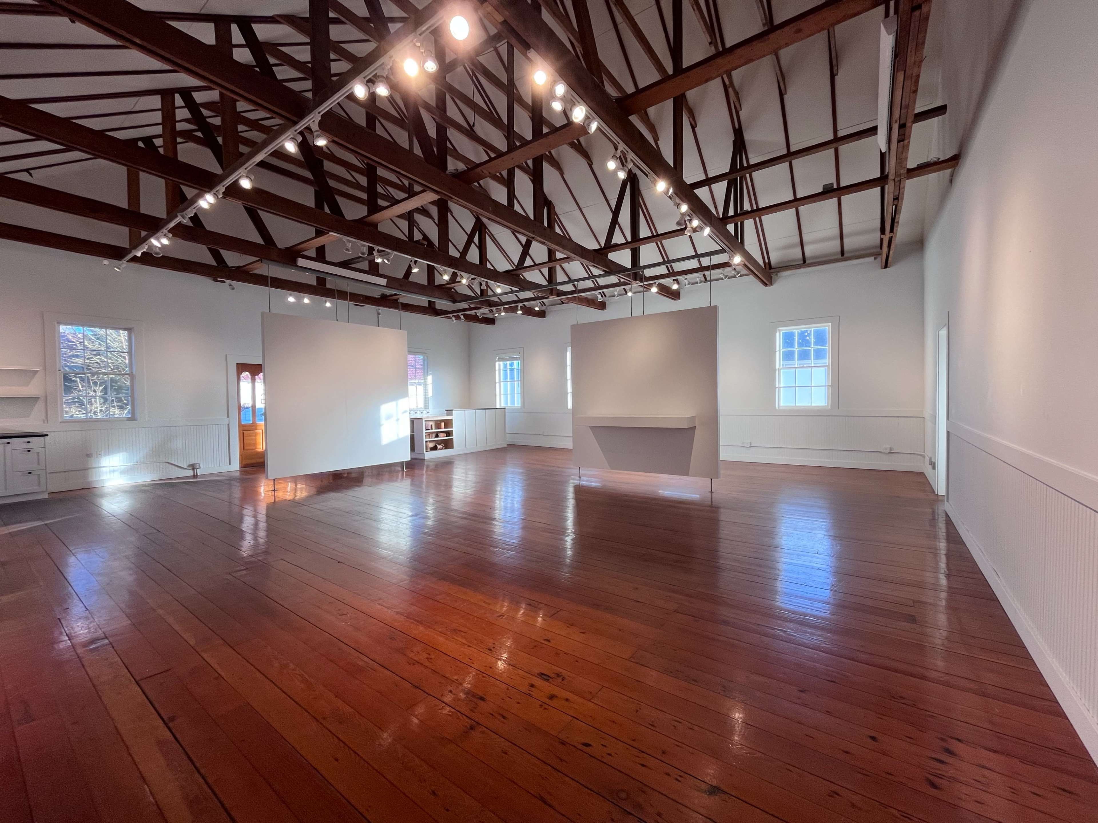 The image shows a spacious interior with polished wooden floors, white walls, and exposed wooden beams, featuring several illuminated display areas.
