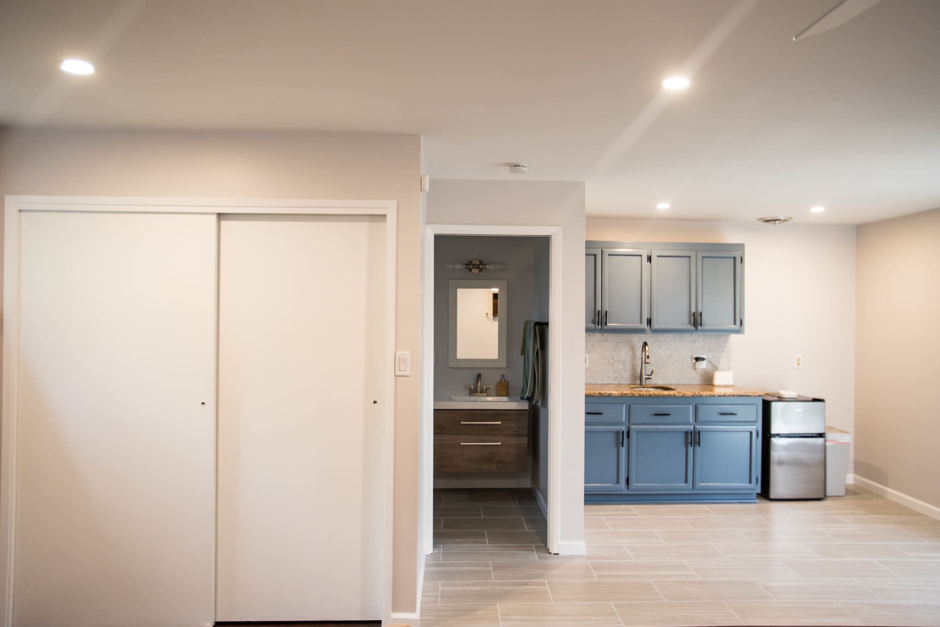 The image shows a light-colored room with a kitchen featuring blue cabinets on the right and a bathroom entrance on the left.
