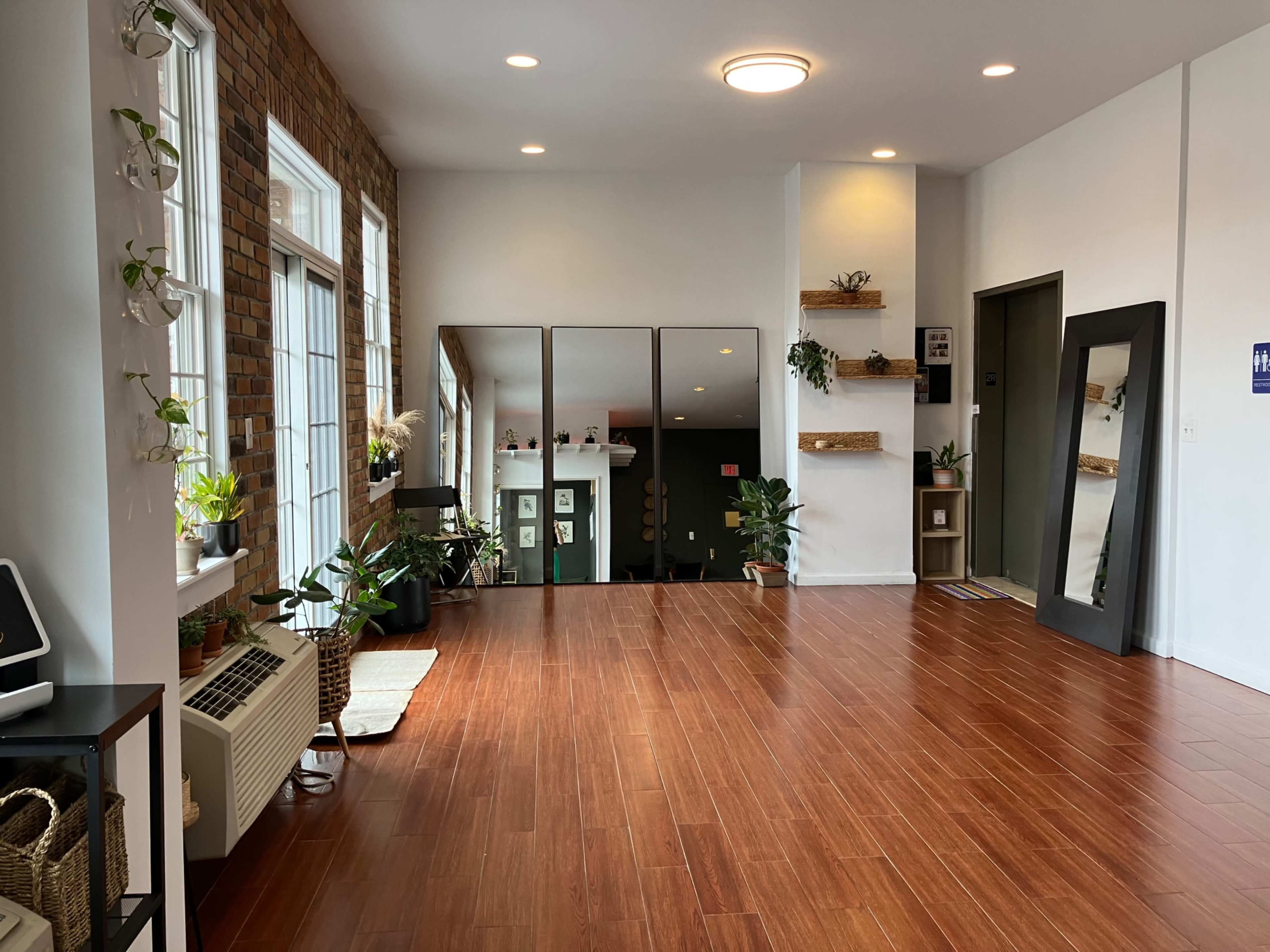 A spacious room with wooden flooring, large windows, and several plants, along with mirrors and an elevator in the background.