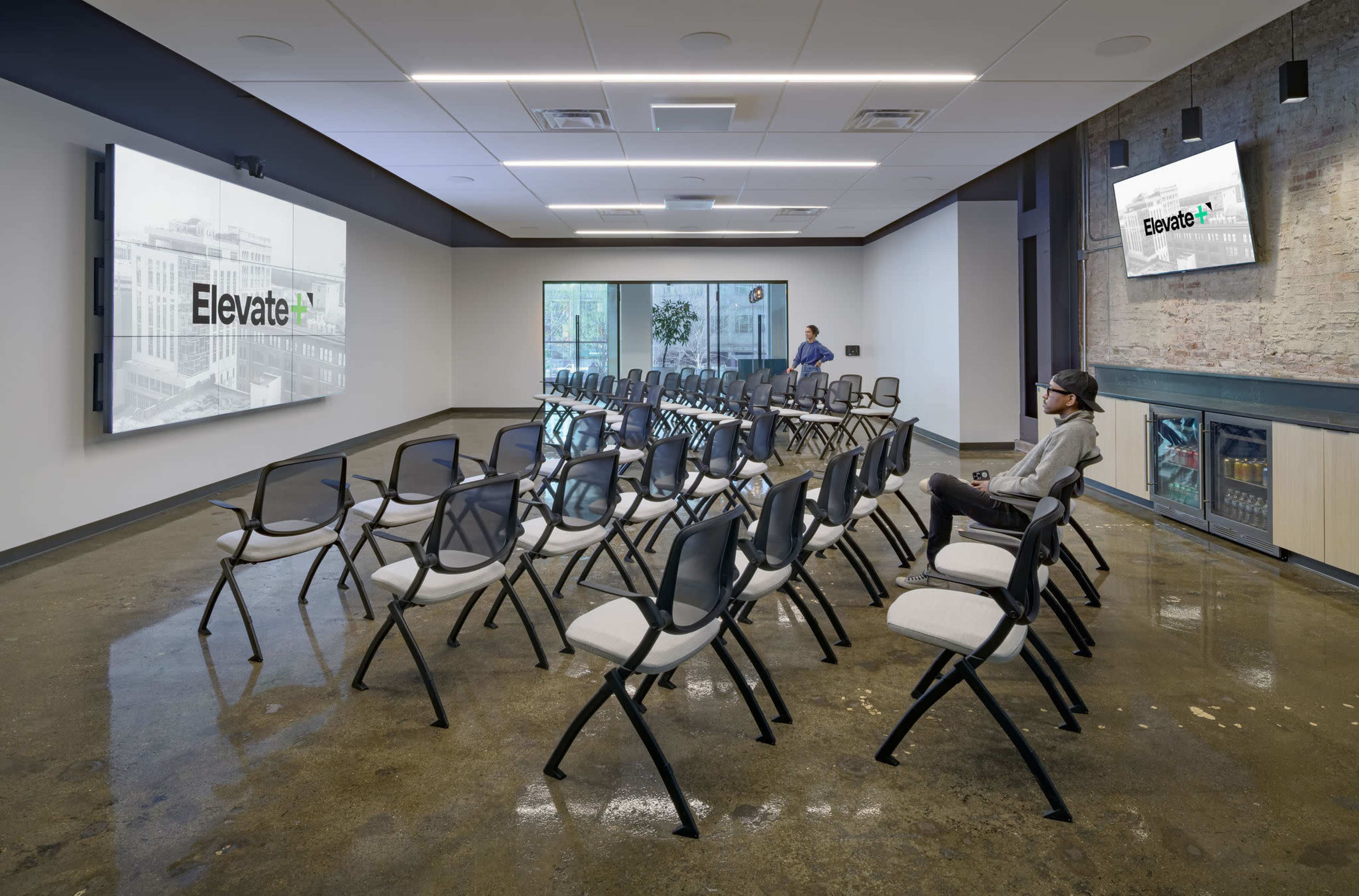 A modern conference room features several rows of black and white folding chairs facing two large screens displaying the word "Elevate."