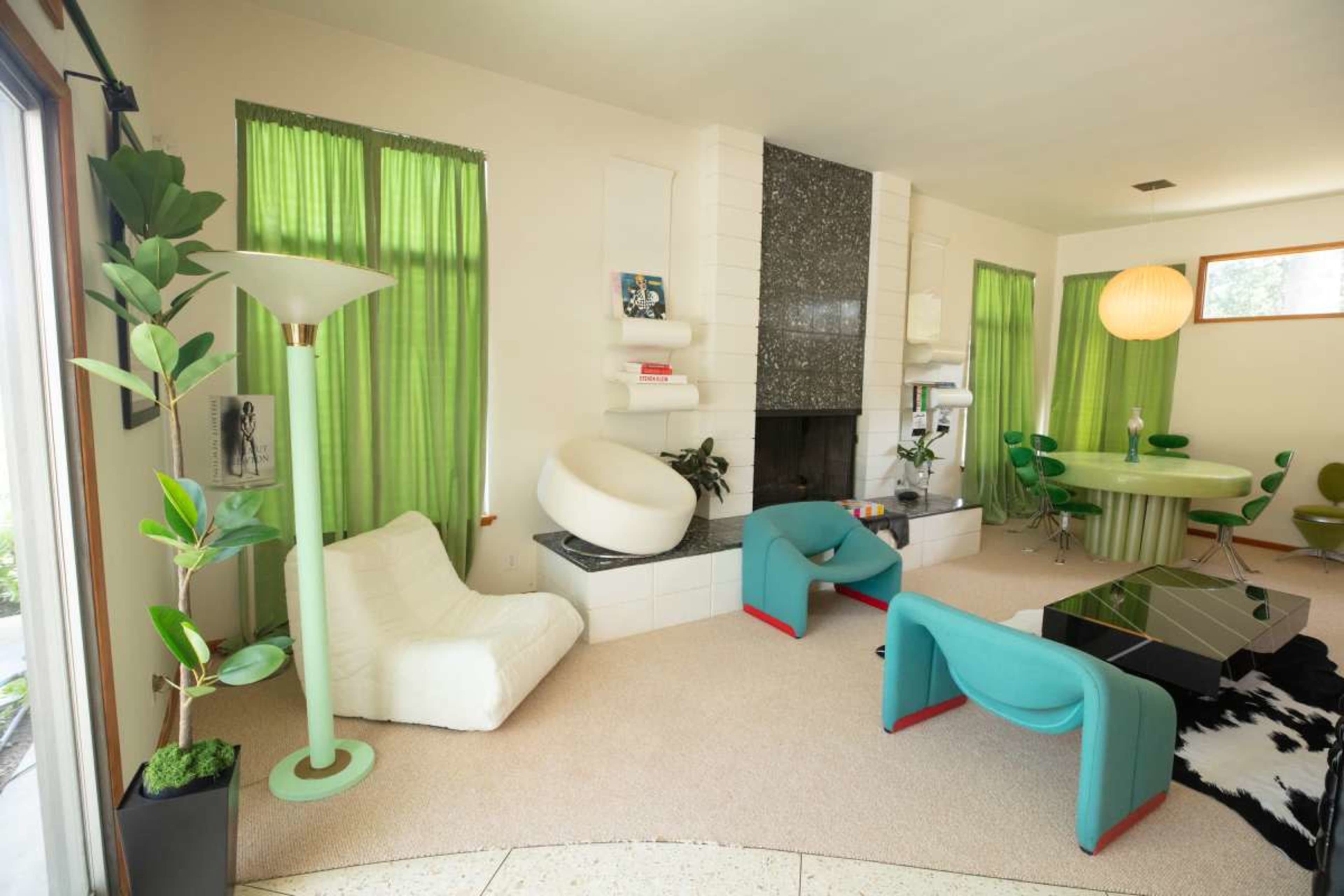 The image features a modern living room with green curtains, a stone fireplace, unique furniture including a circular dining table and colorful chairs, and a cozy seating area with a bean bag and cushions.