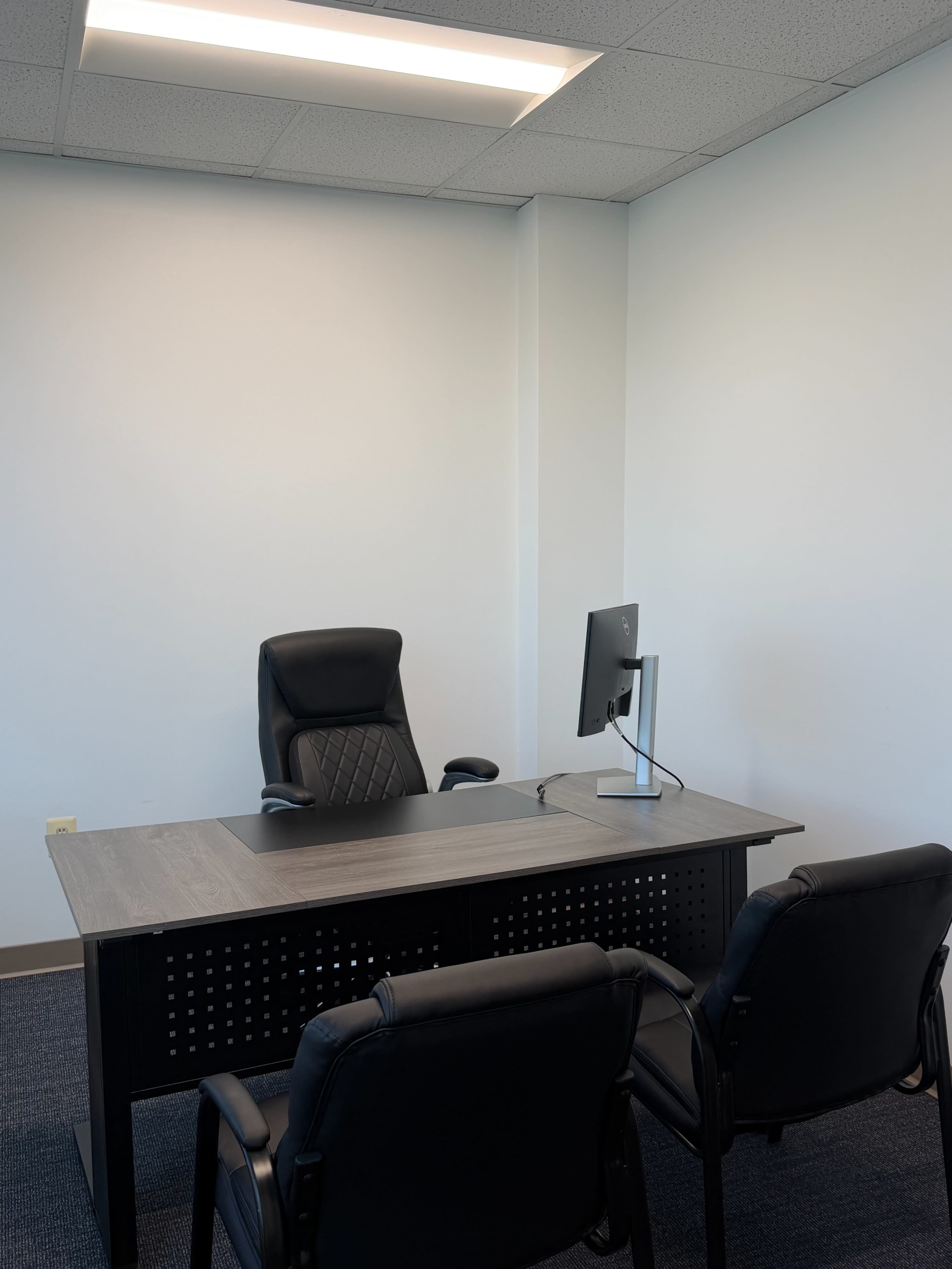 The image shows a simple office setup with a desk, a chair, and a computer monitor facing two black chairs.